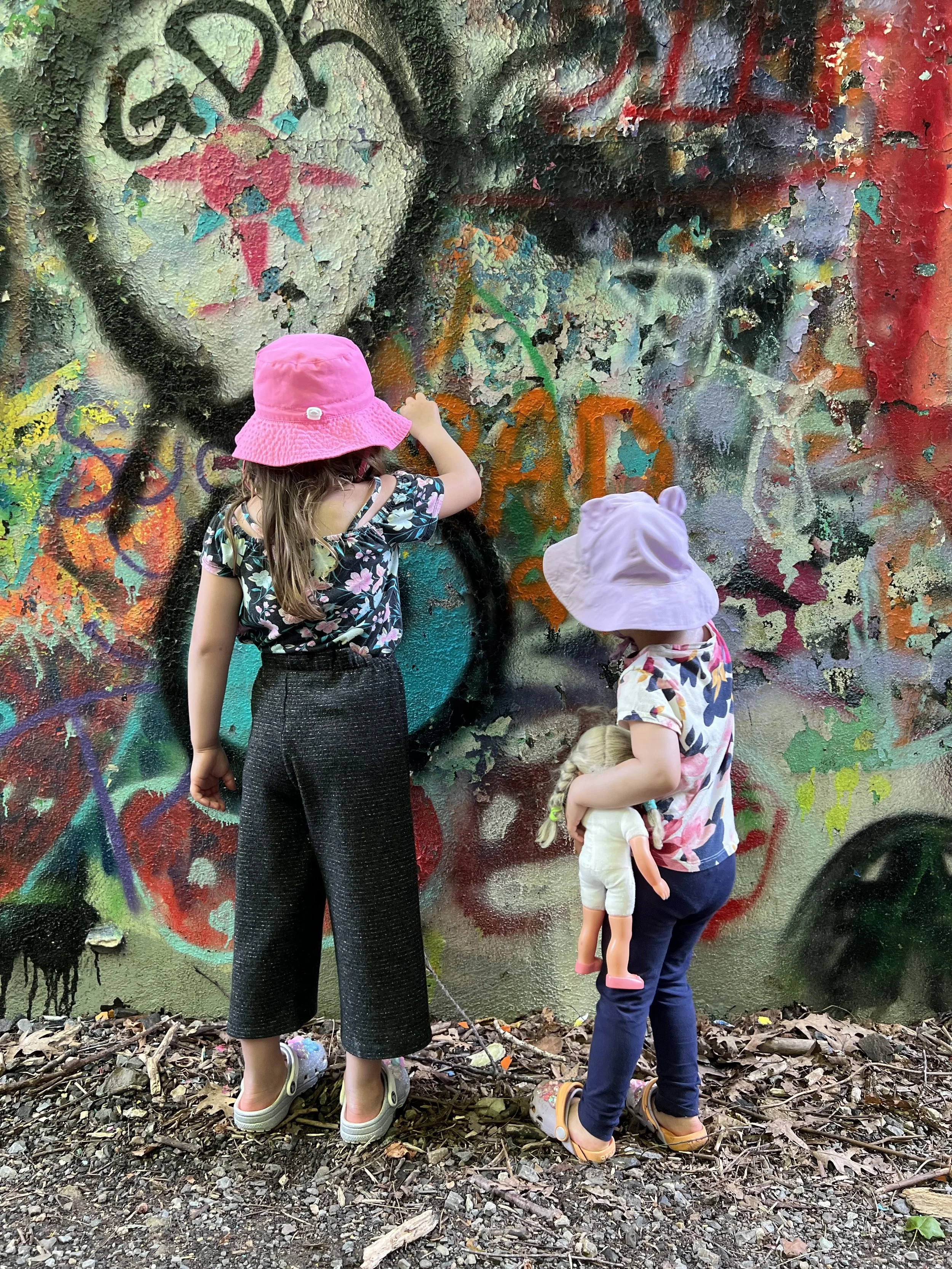 Two young girls wearing pink hats and casual clothes, one holding a doll, stand in front of a colorful graffiti wall. The graffiti features a large heart with a face, surrounded by various abstract shapes and patterns.