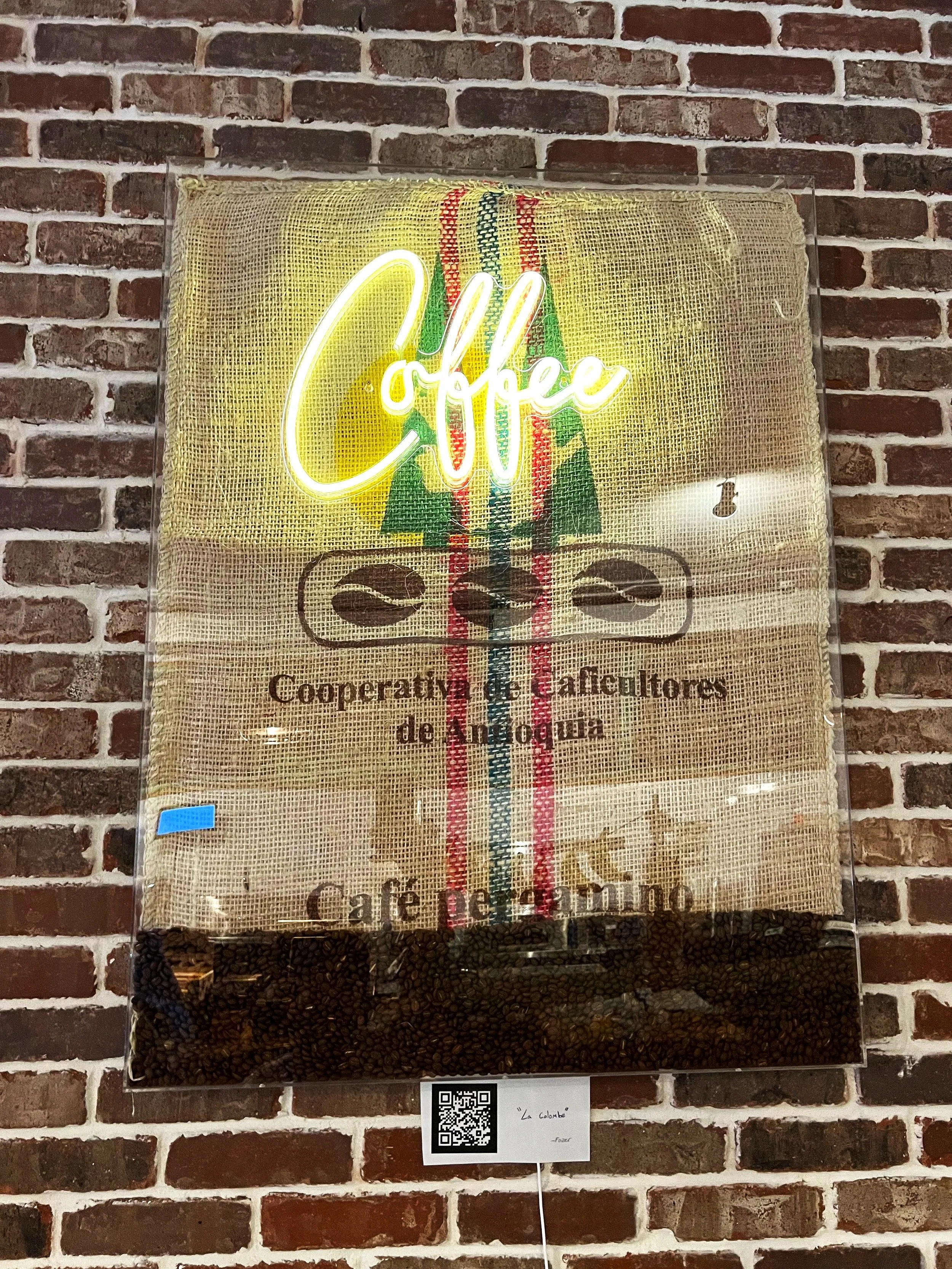 Neon sign reading 'Coffee' placed on a burlap background with coffee beans along the bottom, mounted on a brick wall.