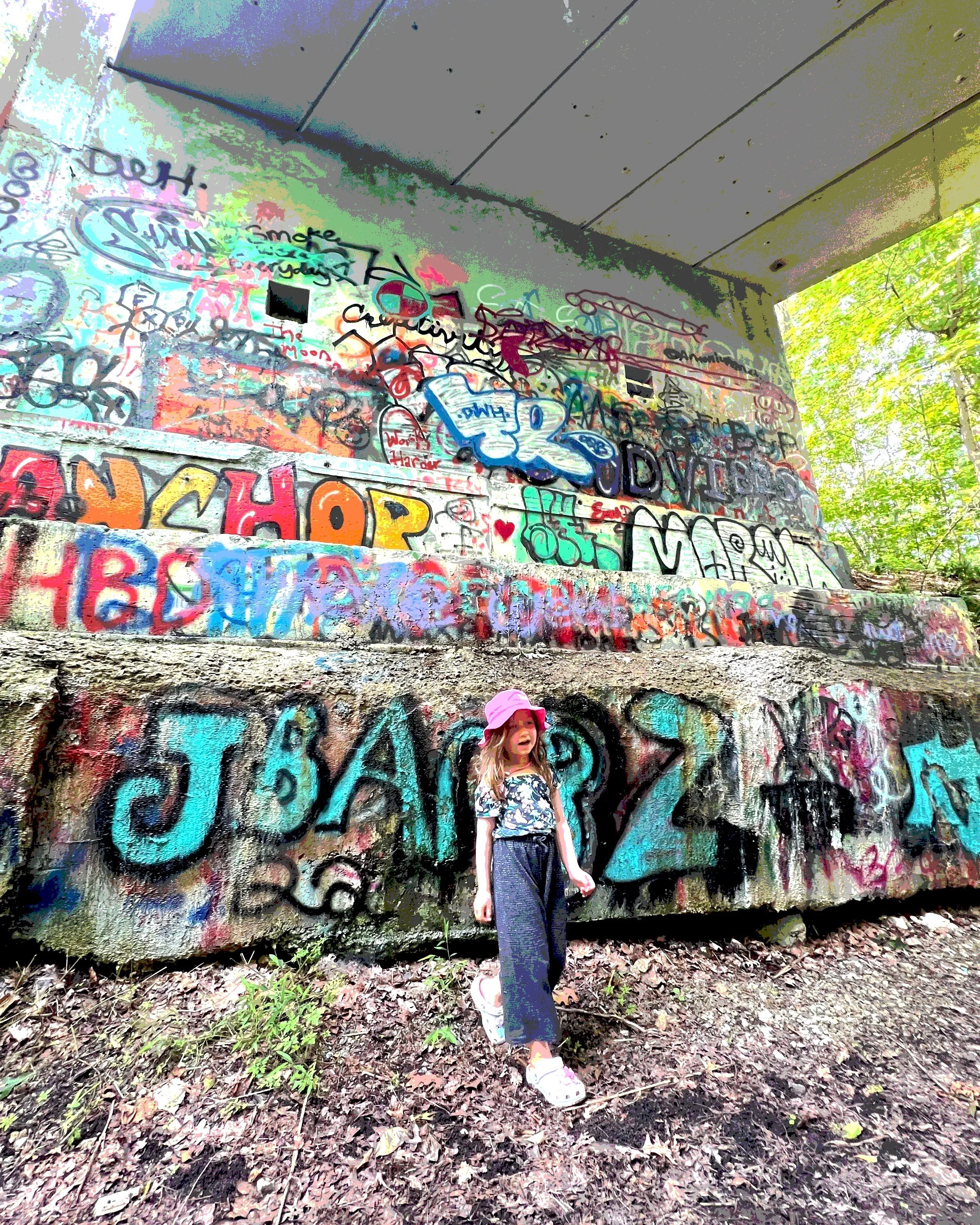 A young girl wearing a pink hat, floral shirt, and dark pants stands in front of a wall covered in colorful graffiti and street art under a bridge, with trees visible on the right side.