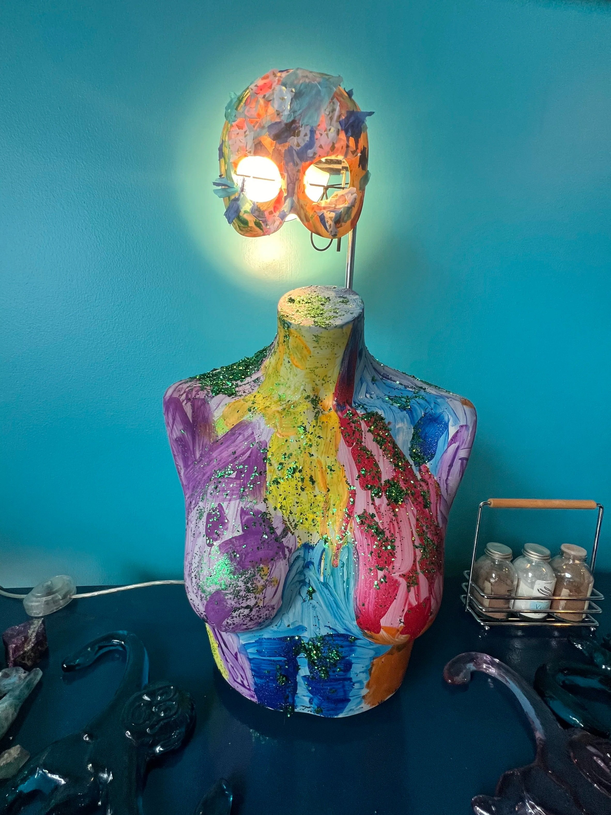 Colorful abstract sculpture of a human torso with a mask-shaped light fixture on top, decorated with paint and glitter, against a blue wall.