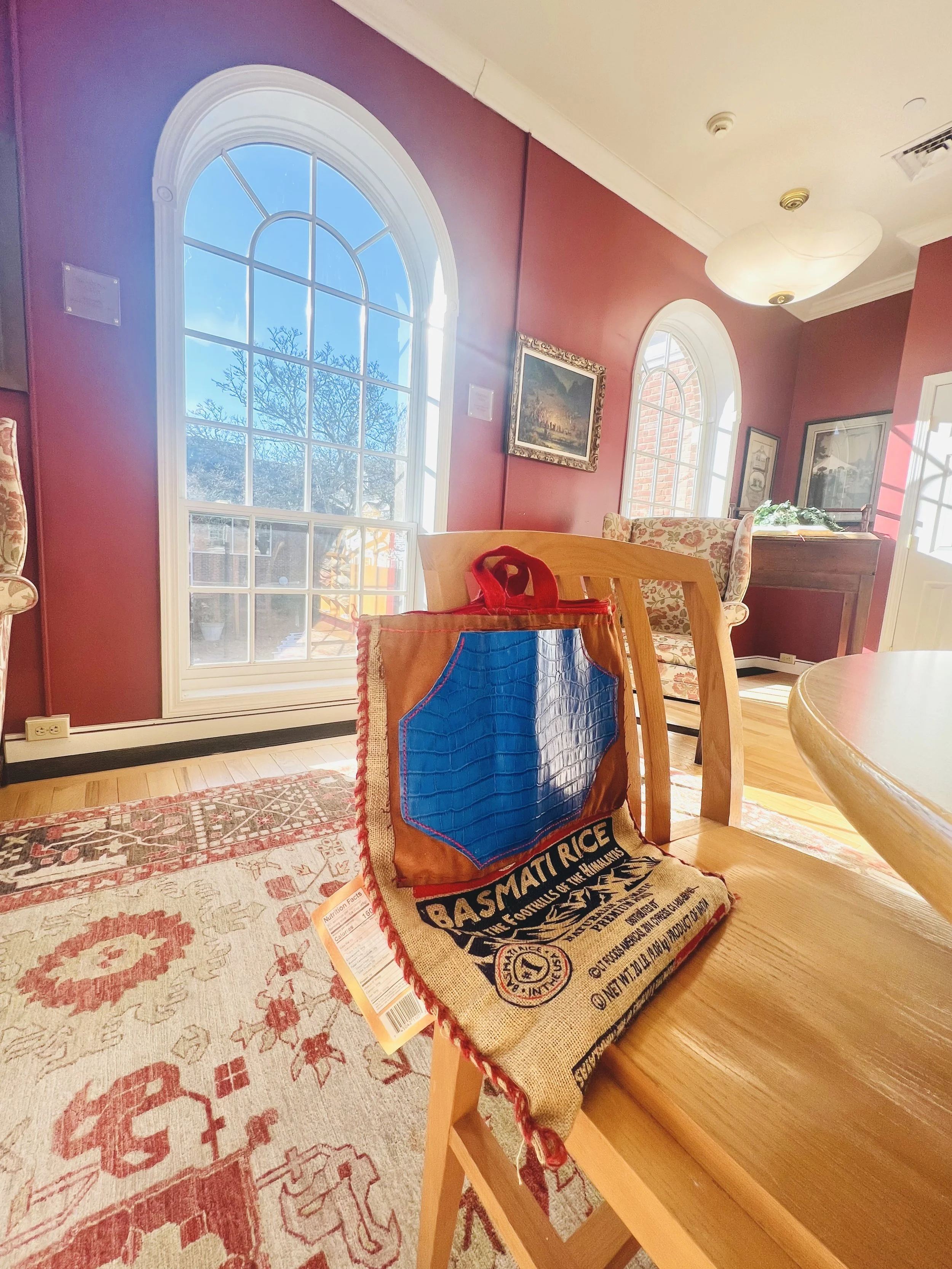 A cozy living room with large arched windows, red walls, and floral armchairs. A colorful bag with rice branding rests on a wooden dining chair.