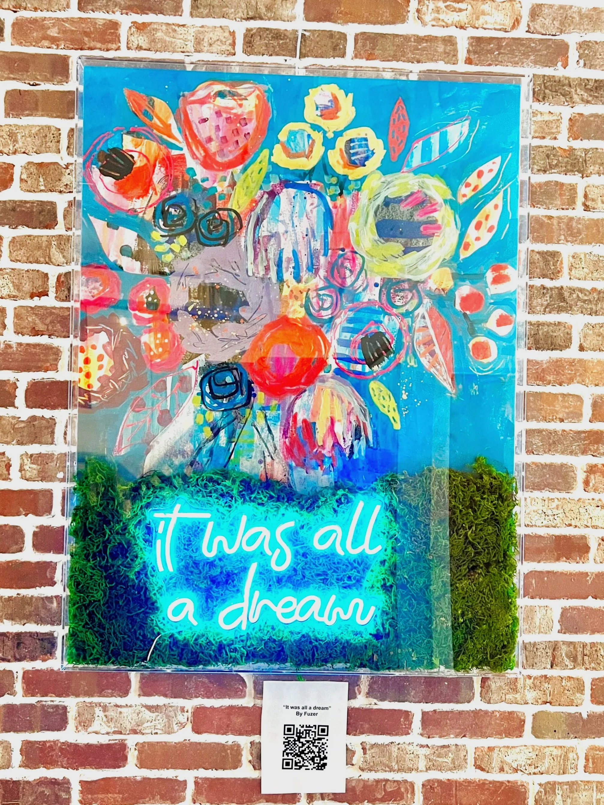 Bright, colorful abstract floral artwork on a blue background with glowing neon text that reads 'it was all a dream', mounted on a brick wall.