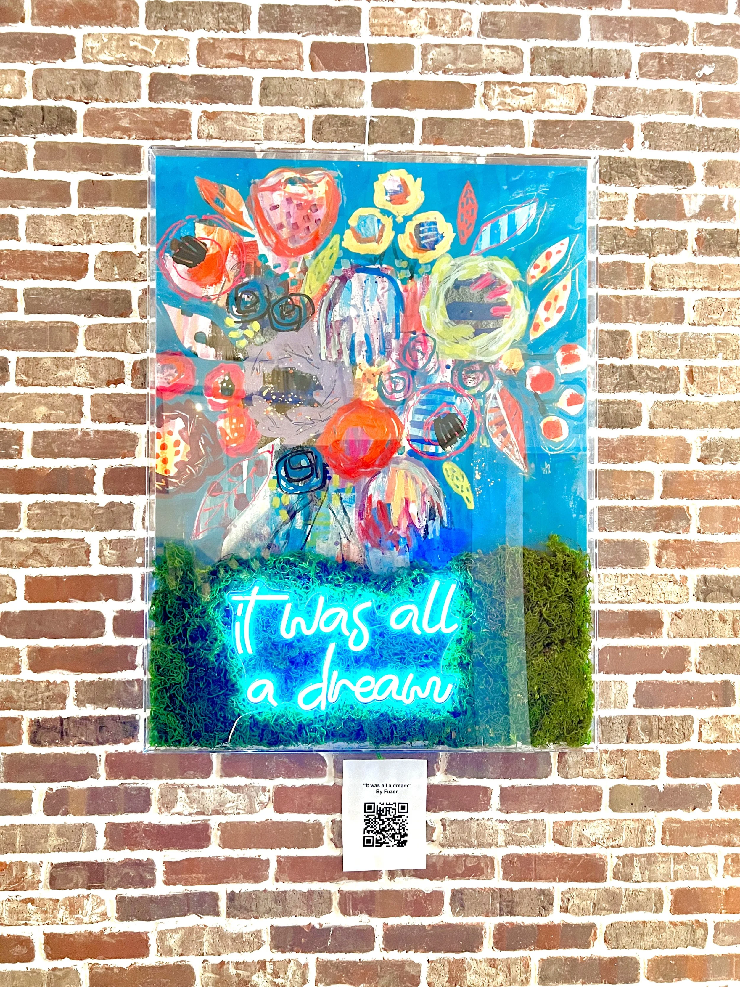 Colorful abstract painting of flowers on a blue background with a neon sign at the bottom that reads 'it was all a dream', mounted on a brick wall.