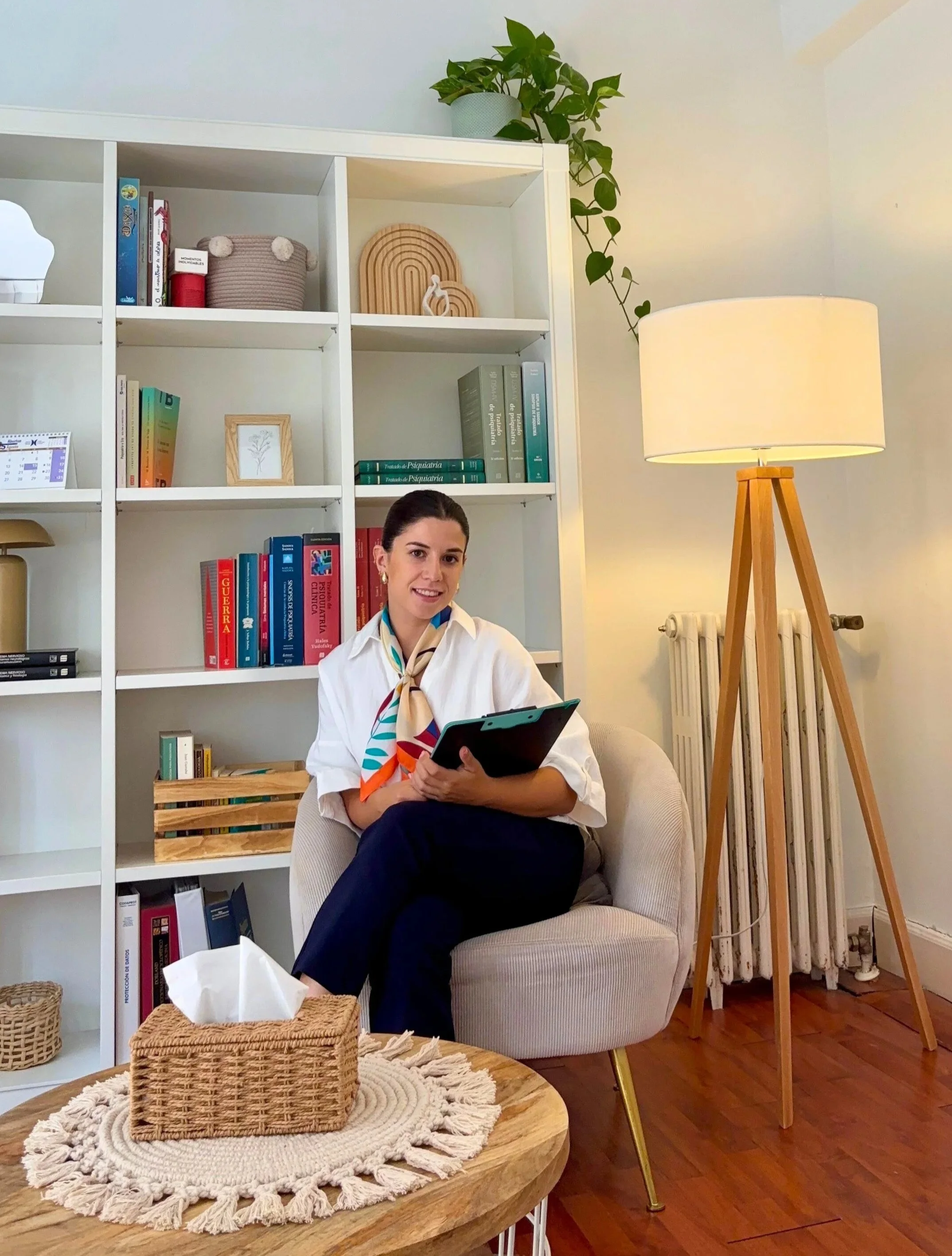 Mujer sentada en una sala, sosteniendo una tableta, con estantería y lámpara de pie en el fondo.