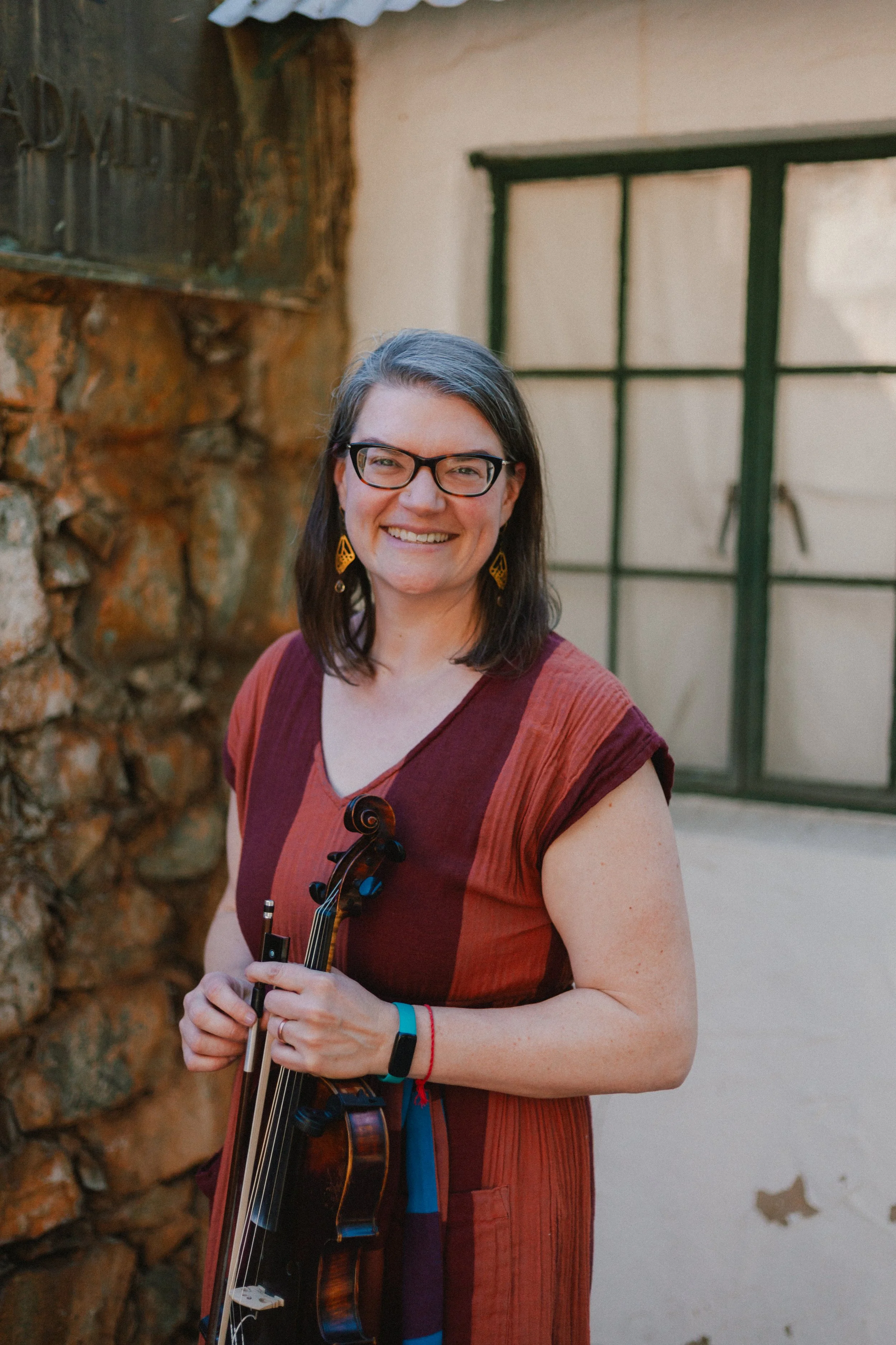 A woman with glasses and earrings smiling and holding a violin, standing outdoors with a stone wall and window in the background.