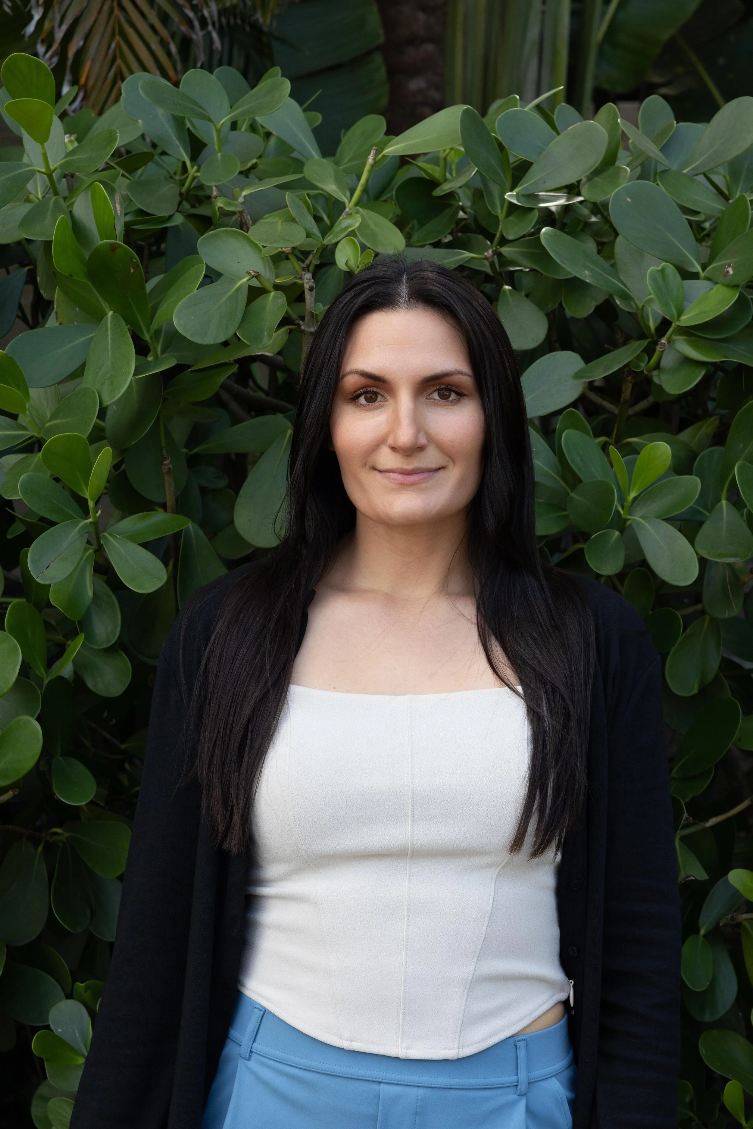 A woman with long dark hair dressed in a white top and blue pants standing in front of green leafy foliage.