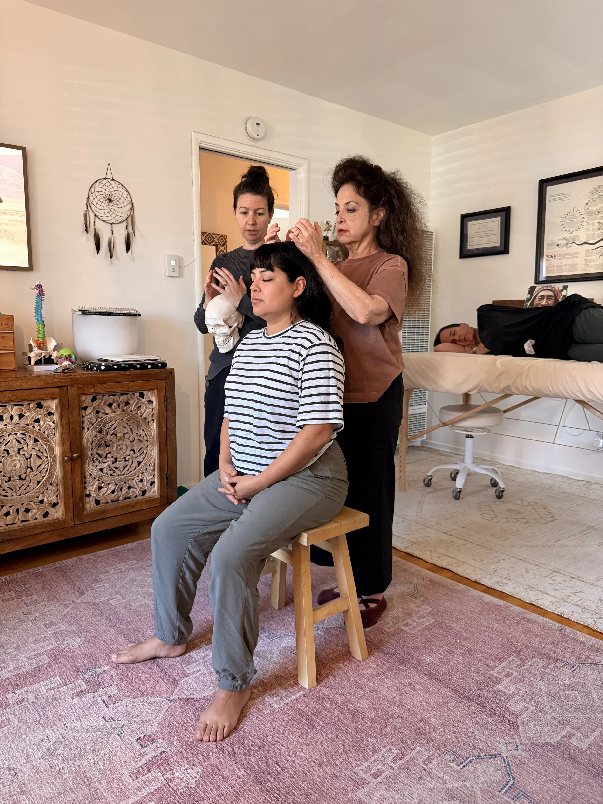 A woman is sitting on a stool with her eyes closed, receiving a spiritual somatic healing session from a student standing behind her while her teacher shows her the technique in a room with a massage table and wall decorations.