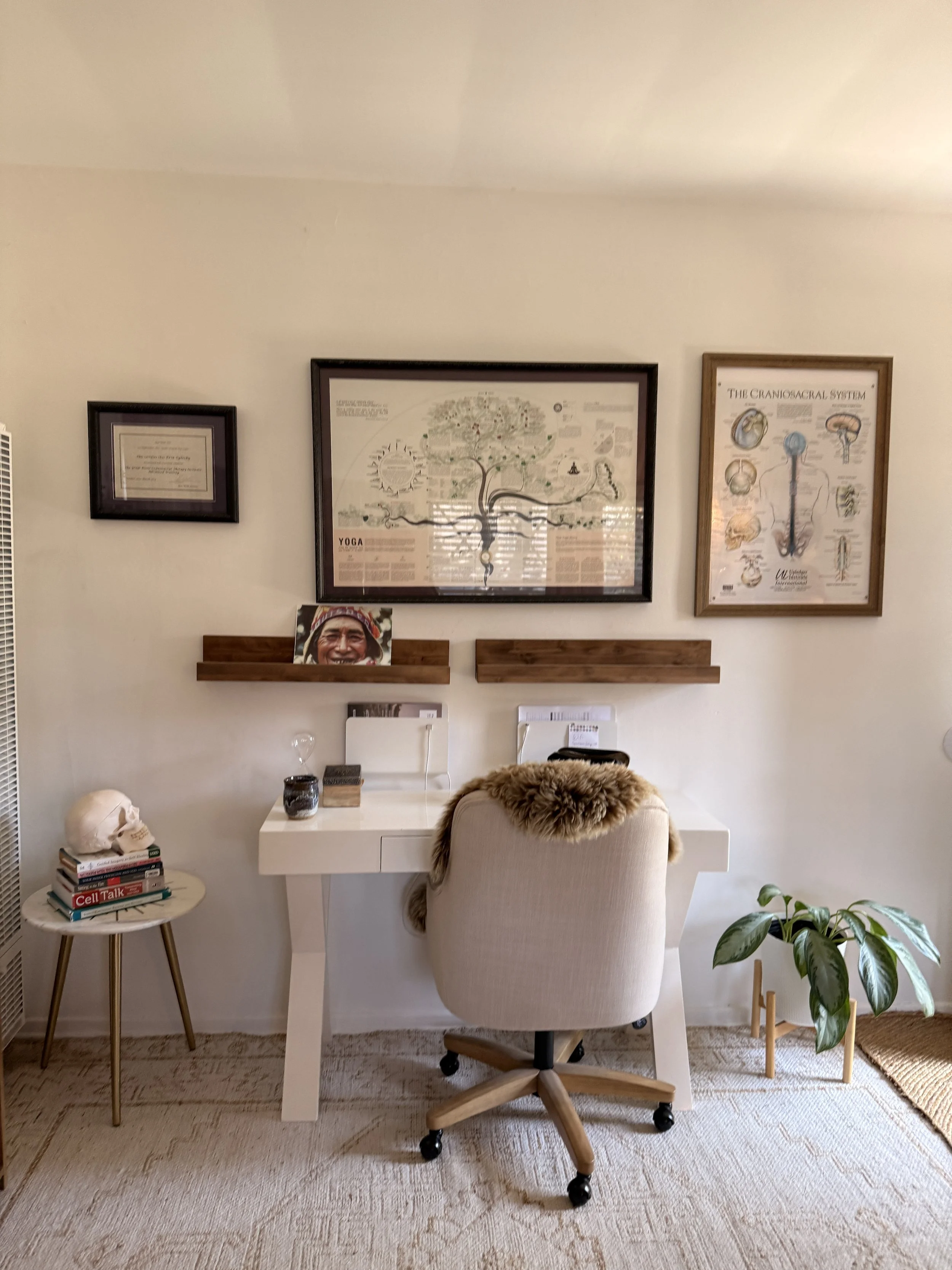 Home office for remote healing sessions with a white desk, a beige swivel chair with a fur throw, potted plant, bookshelf, framed art and posters on the wall, and a small side table with craniosacral healing books and a skull replica.