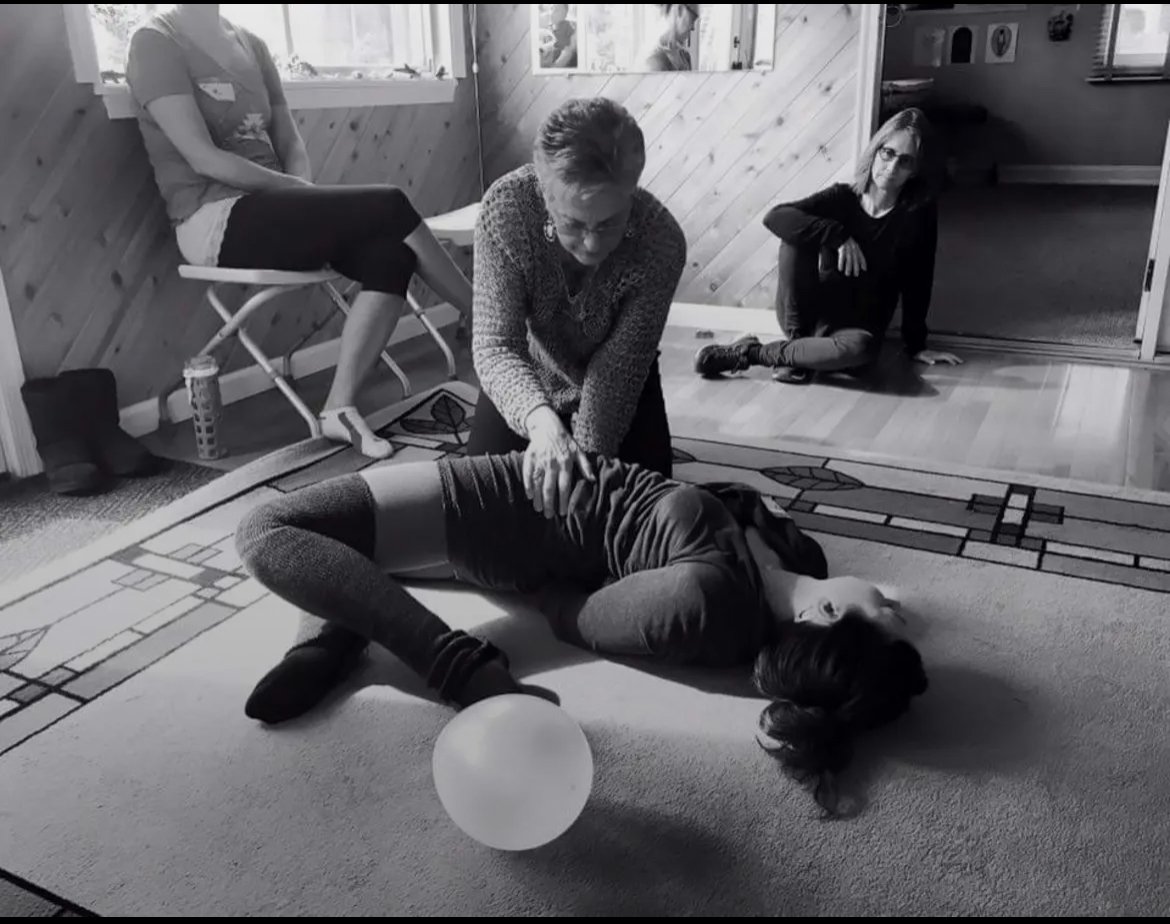 A woman is performing CPR on a person lying on the floor, while two other women observe. One woman is sitting on a chair, and the other is sitting on the floor near the doorway.