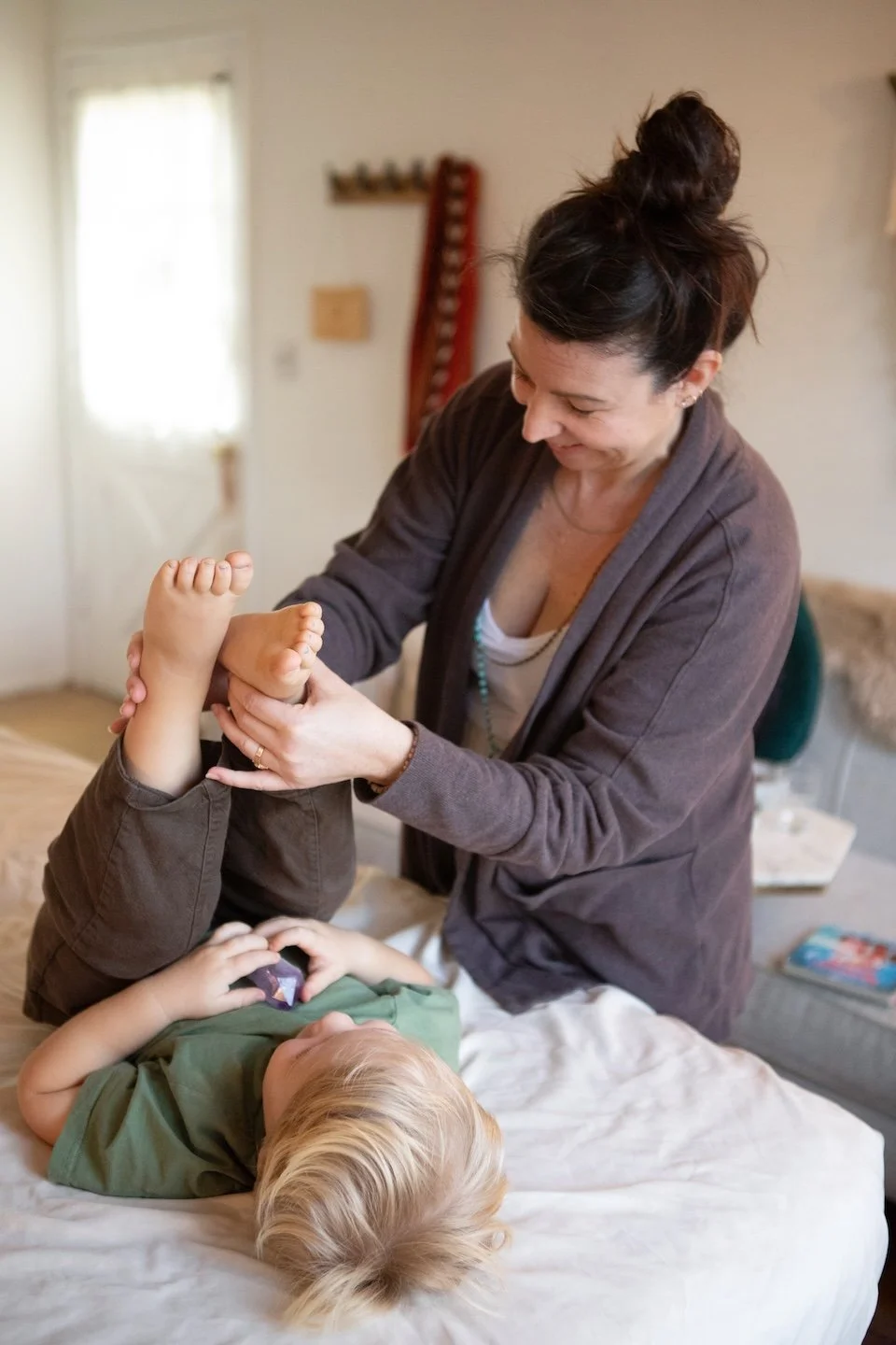 A woman is standing over a child lying on a bed, holding the child's feet and smiling, in a cozy bedroom with natural light.