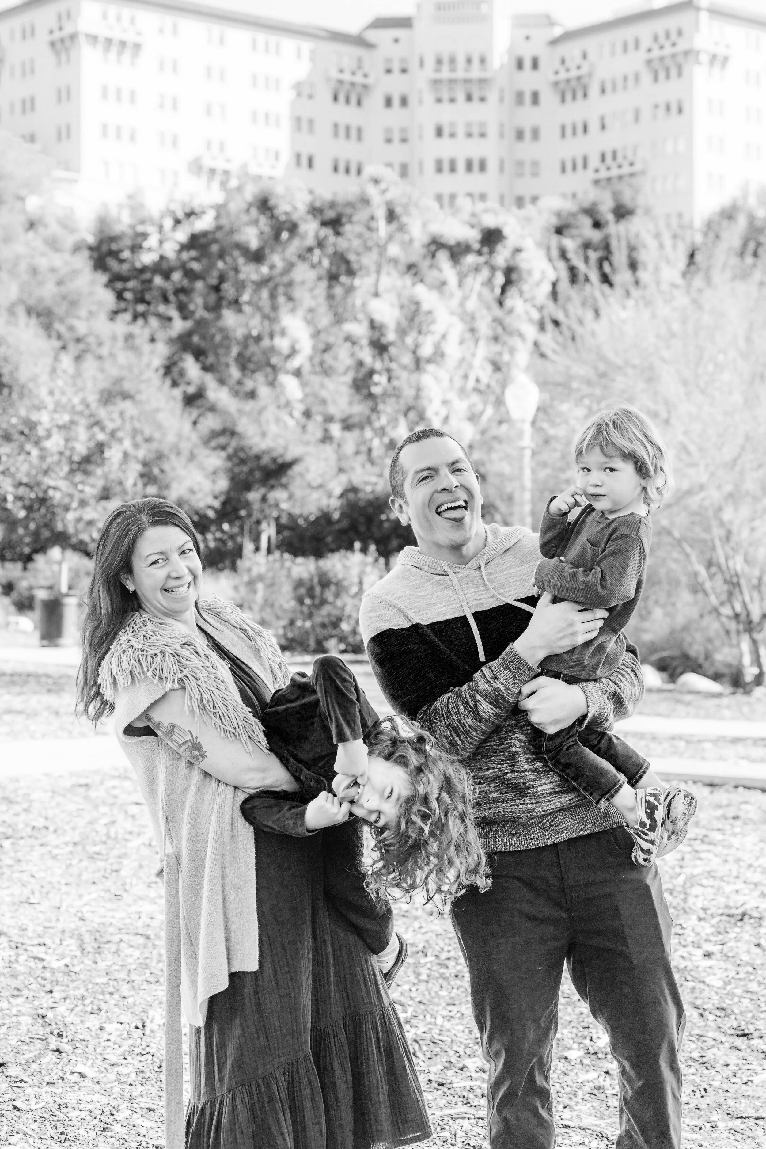 A black and white photo of a family of four outdoors. The woman is holding a boy, while the man is holding a girl by the waist. They are all smiling and laughing.