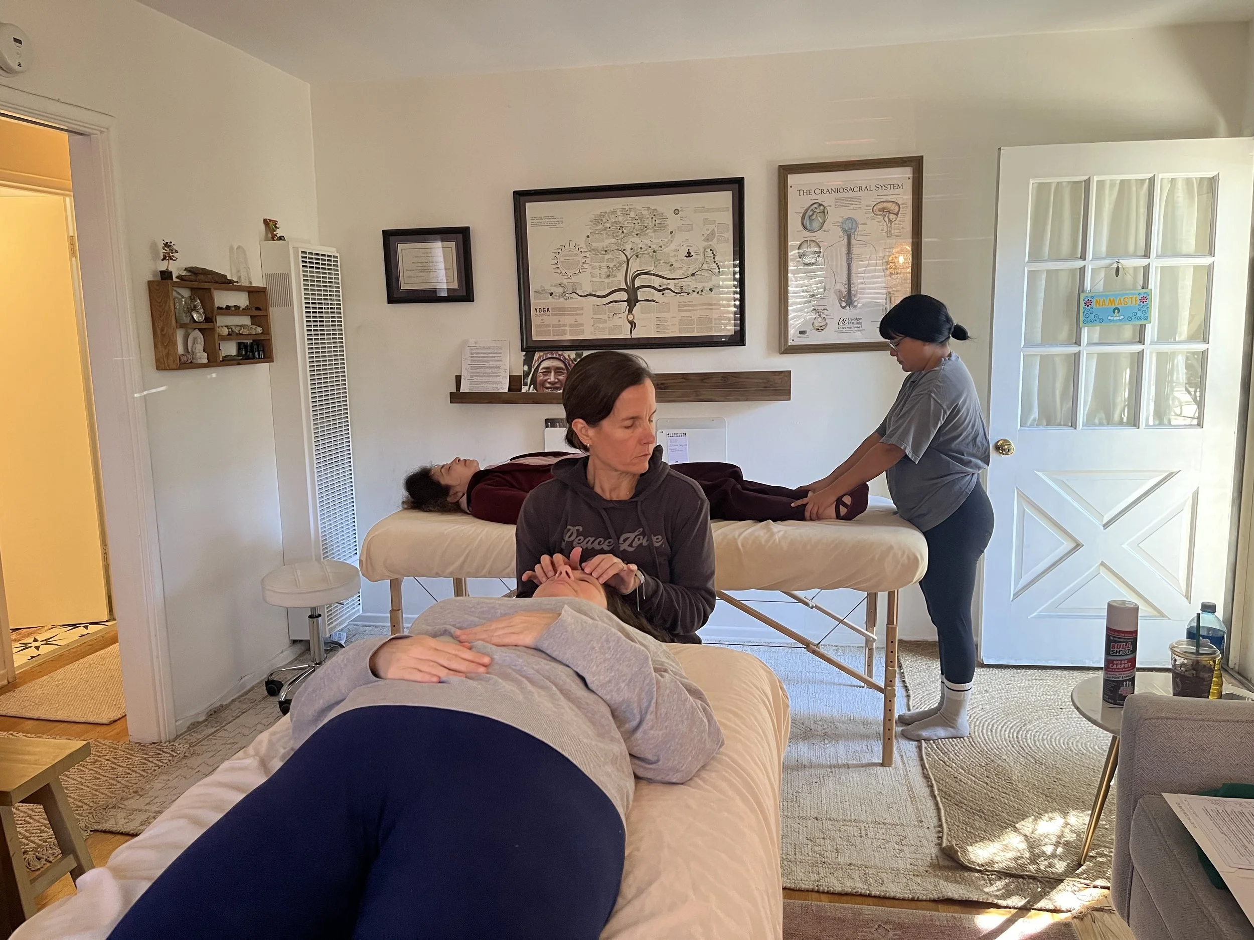 Two women receiving massages in a cozy, well-lit room with framed posters on the wall and natural light coming through a glass-paneled door.