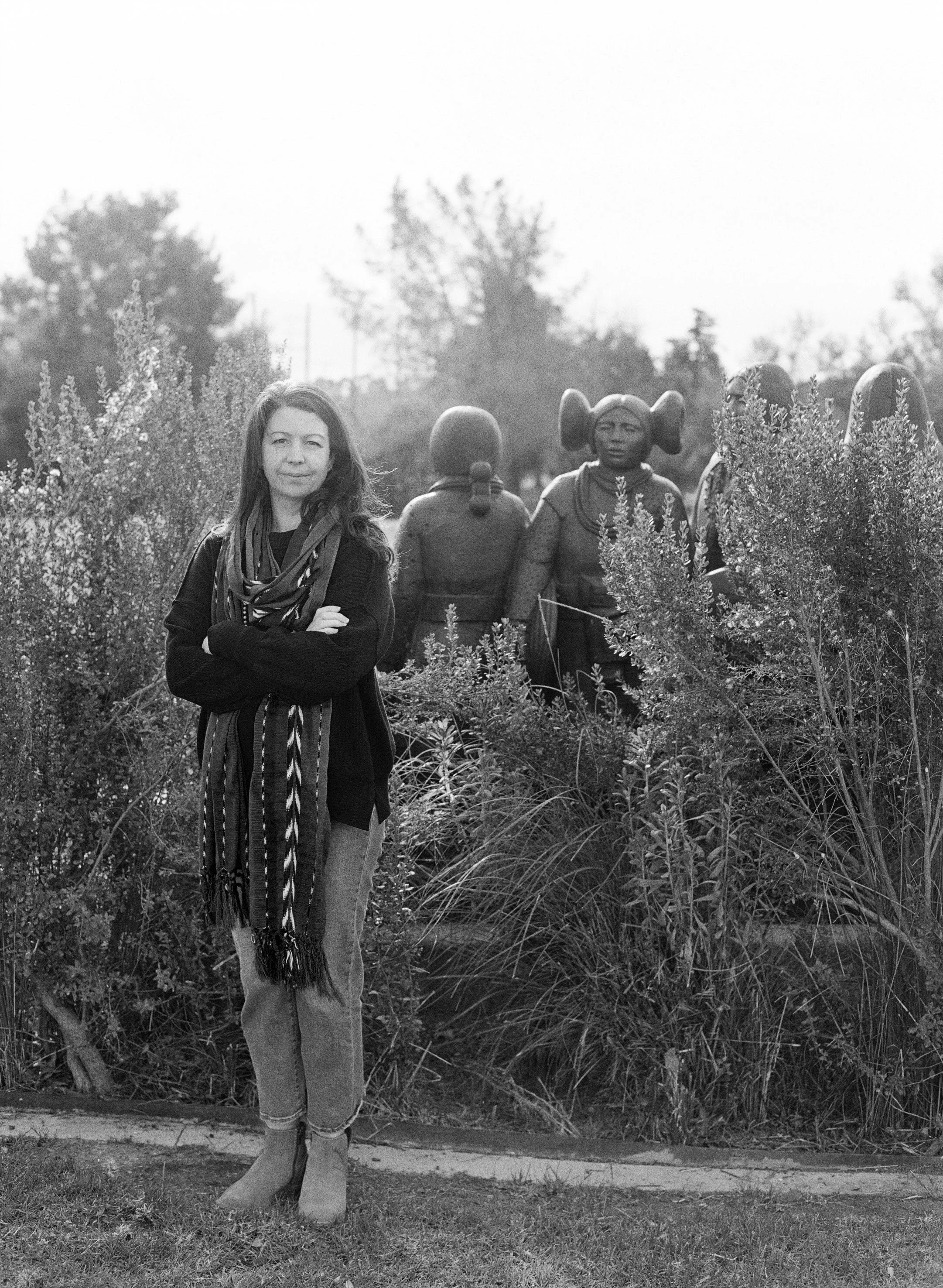 A woman with long hair, wearing a dark jacket and scarf, standing outdoors with her arms crossed. In the background, there are sculptures of children and plants.