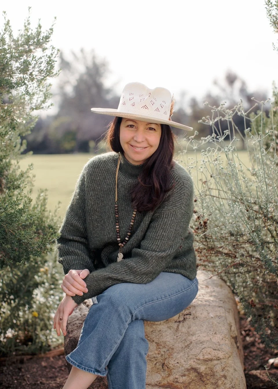 Erin wearing a wide-brimmed straw hat, a green sweater, and blue jeans sitting on a large rock outdoors among greenery with a blurred background.