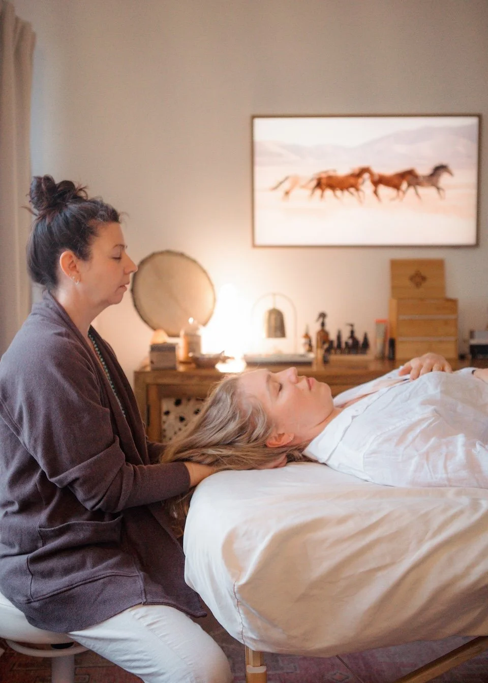 A woman lying in bed with her eyes closed, receiving treatment from Erin. The massage is taking place in a cozy bedroom with warm lighting, a painting of running horses on the wall, and various decorative items on a wooden dresser in the background.
