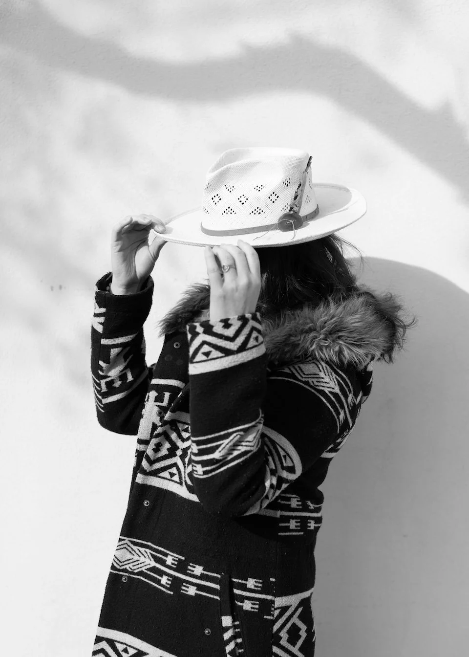 Person wearing a patterned sweater and a fur-lined hood adjusting a straw hat with holes.