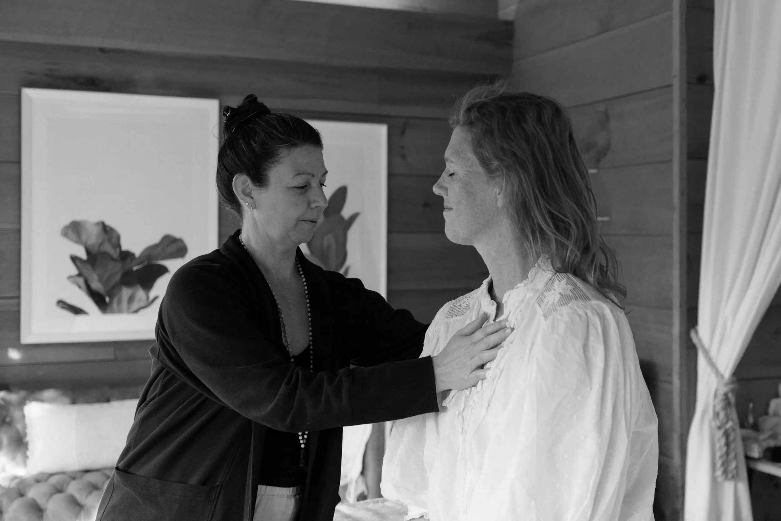A woman with dark hair in a bun places her hand on the chest of another woman with wavy hair, in a room with wood-paneled walls and framed artwork.