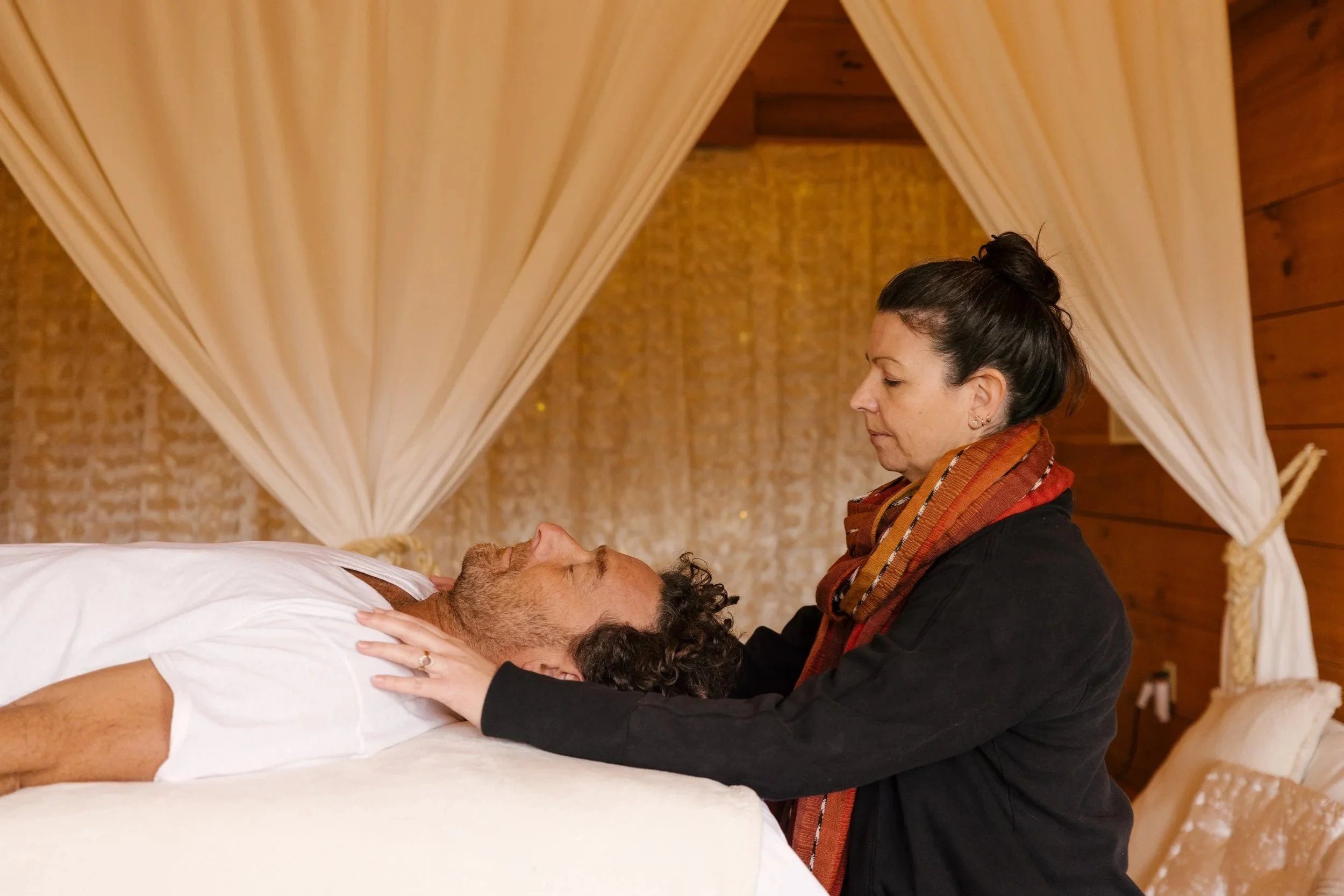 A craniosacral therapist is working on a male client lying on a massage table, holding his head gently and looking at him with a caring expression. The setting is a cozy healing space with warm wooden walls and flowy curtains.
