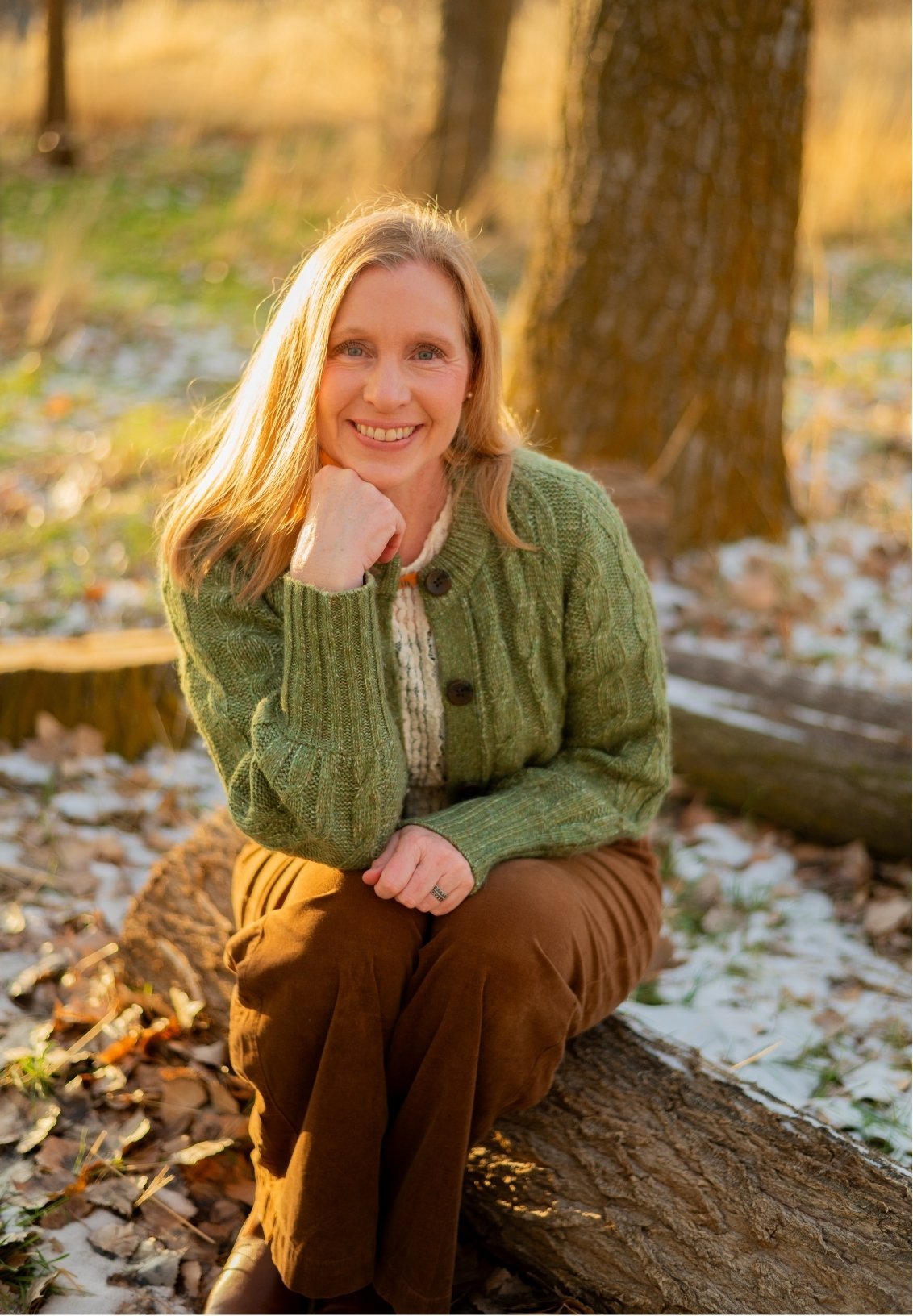 Spanish Fork therapist Kristina Robbins sits on a log in a wooded area during autumn, wearing a green sweater and brown pants, with sunlight filtering through the trees.