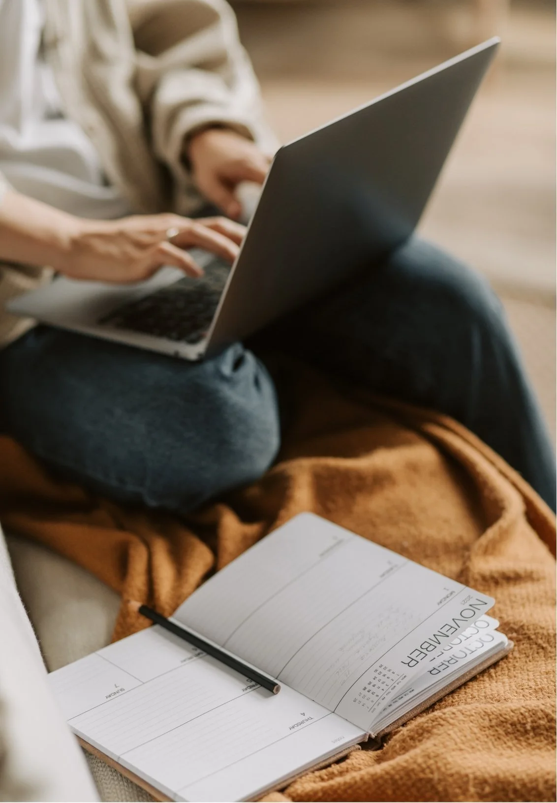 Person working on a laptop with an open planner and pen on a blanket nearby.