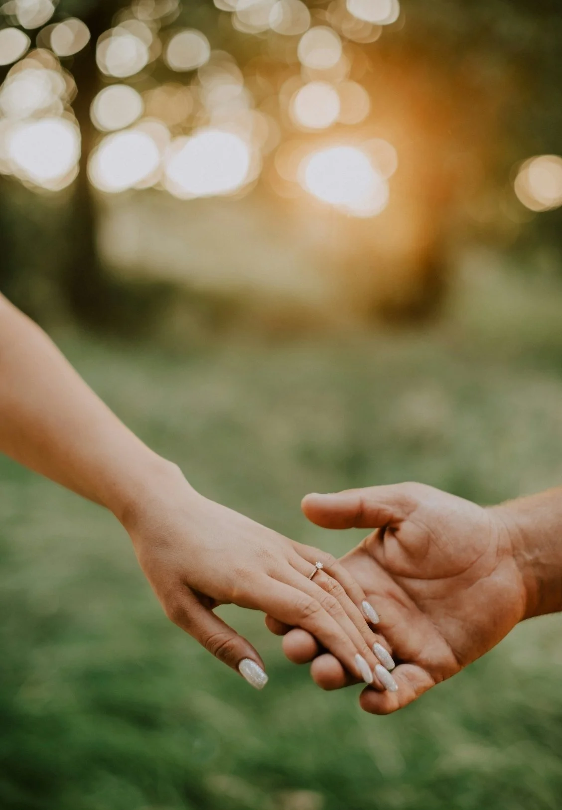 A woman and man holding hands, with the woman's hand showing an engagement ring, outdoors during sunset.