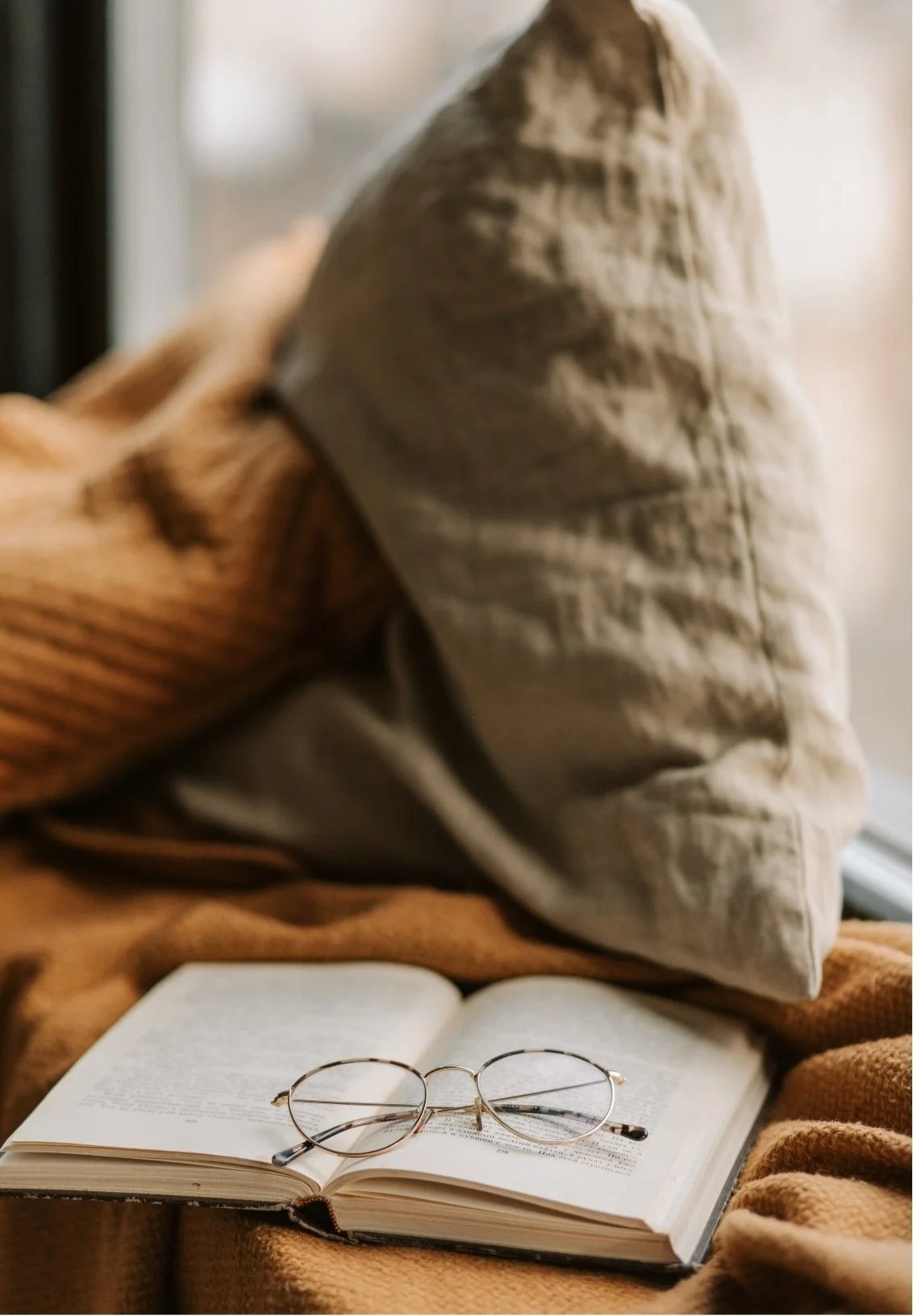 An open book with reading glasses placed on top, placed on a soft brown blanket with a beige pillow nearby, next to a window.