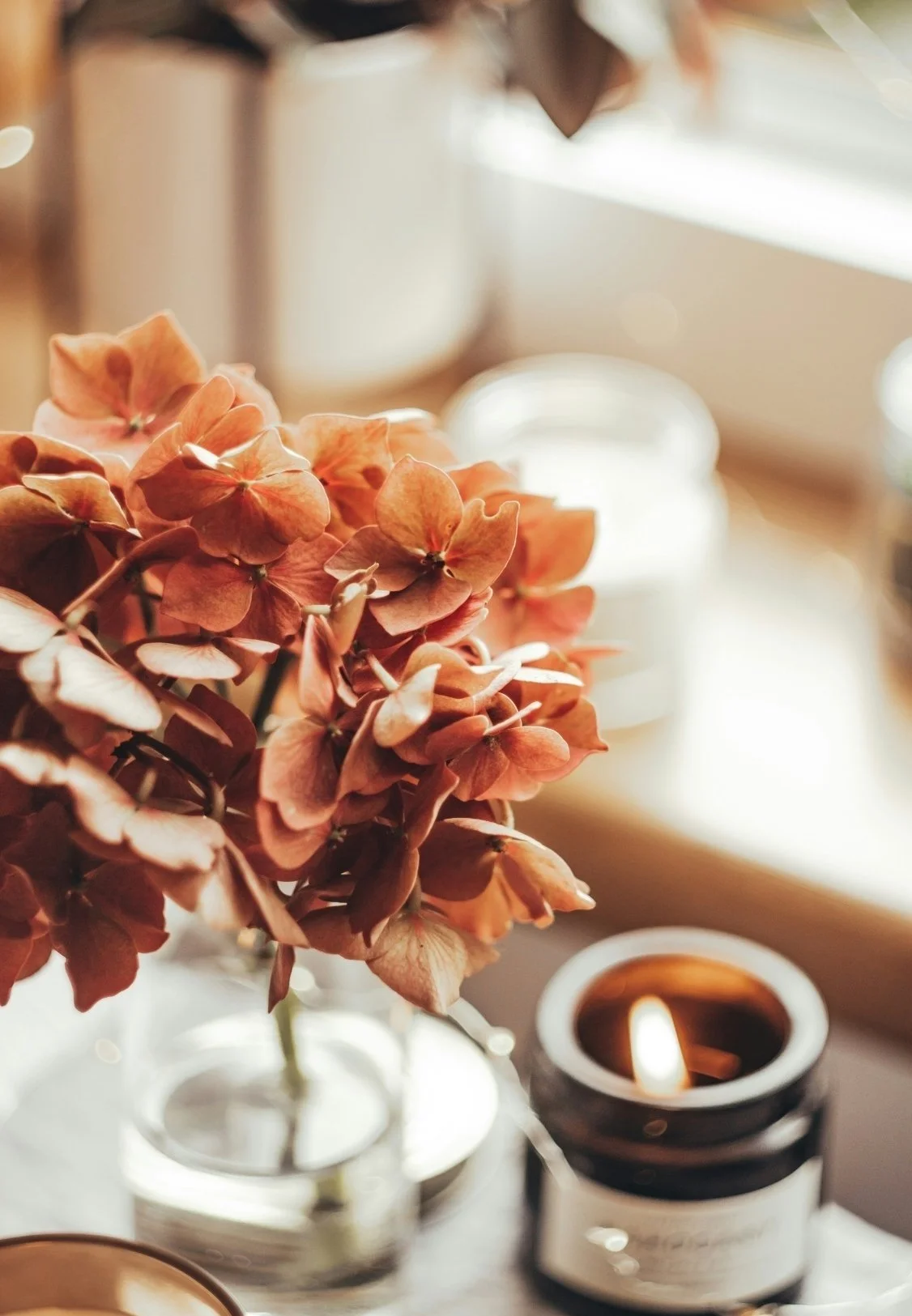 A close-up of a bouquet of dried pink and peach hydrangea flowers in a glass vase, with a lit candle in a black jar and a wooden bowl nearby, on a windowsill with sunlight streaming in.