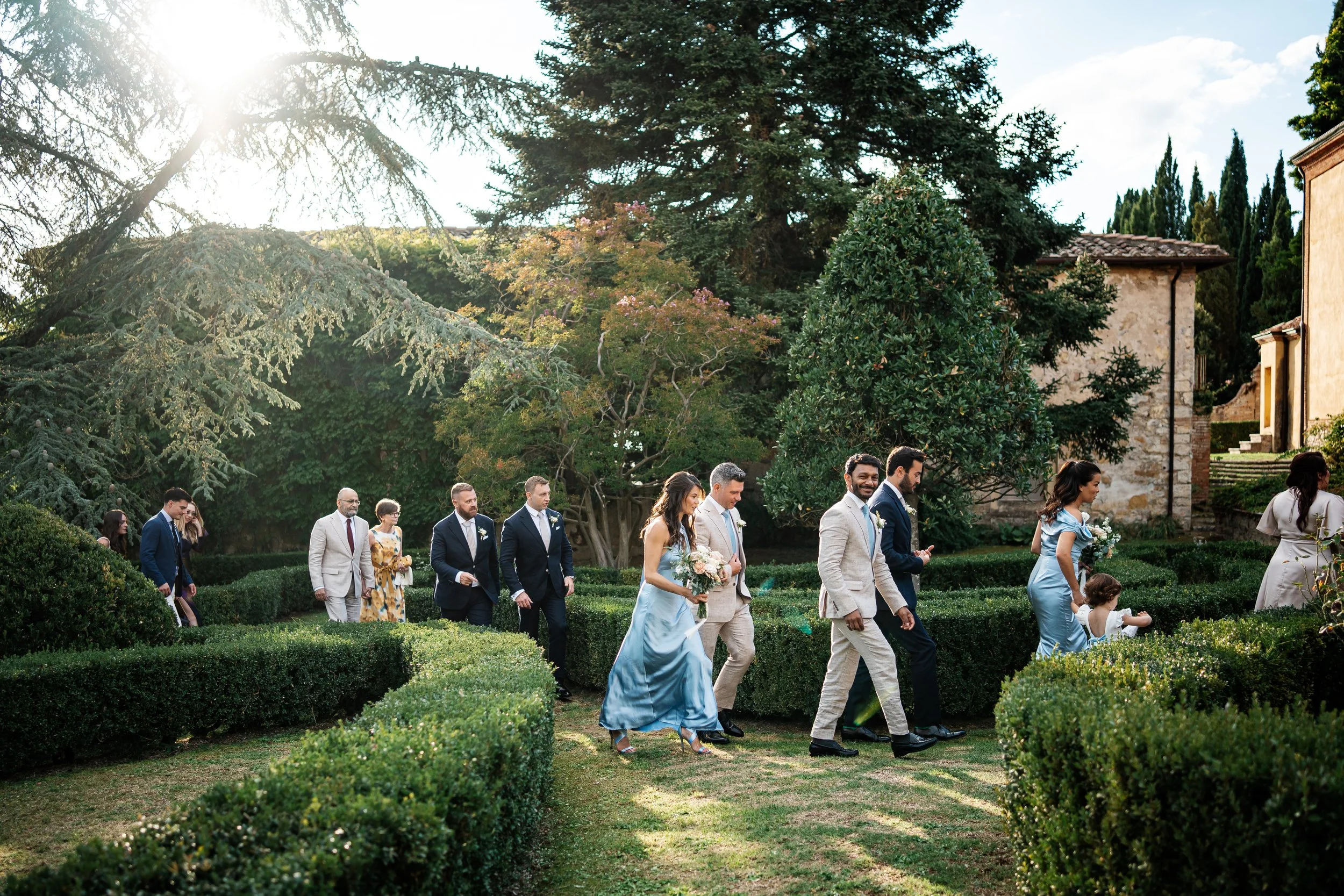 Un gruppo di persone partecipa a un matrimonio all'aperto in un giardino, con uomini in giacca e donne in abiti eleganti, camminando lungo un percorso tra siepi.