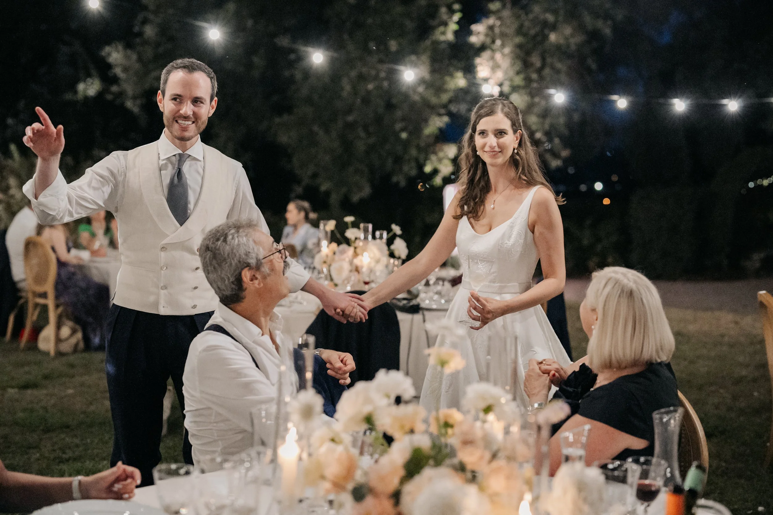 Una coppia di sposi sul tavolo degli invitati durante un matrimonio, con il marito che tiene la mano della moglie e un uomo più anziano seduto che li guarda sorridendo, di sera con luci decorative sopra e tavoli decorati con fiori e candele.