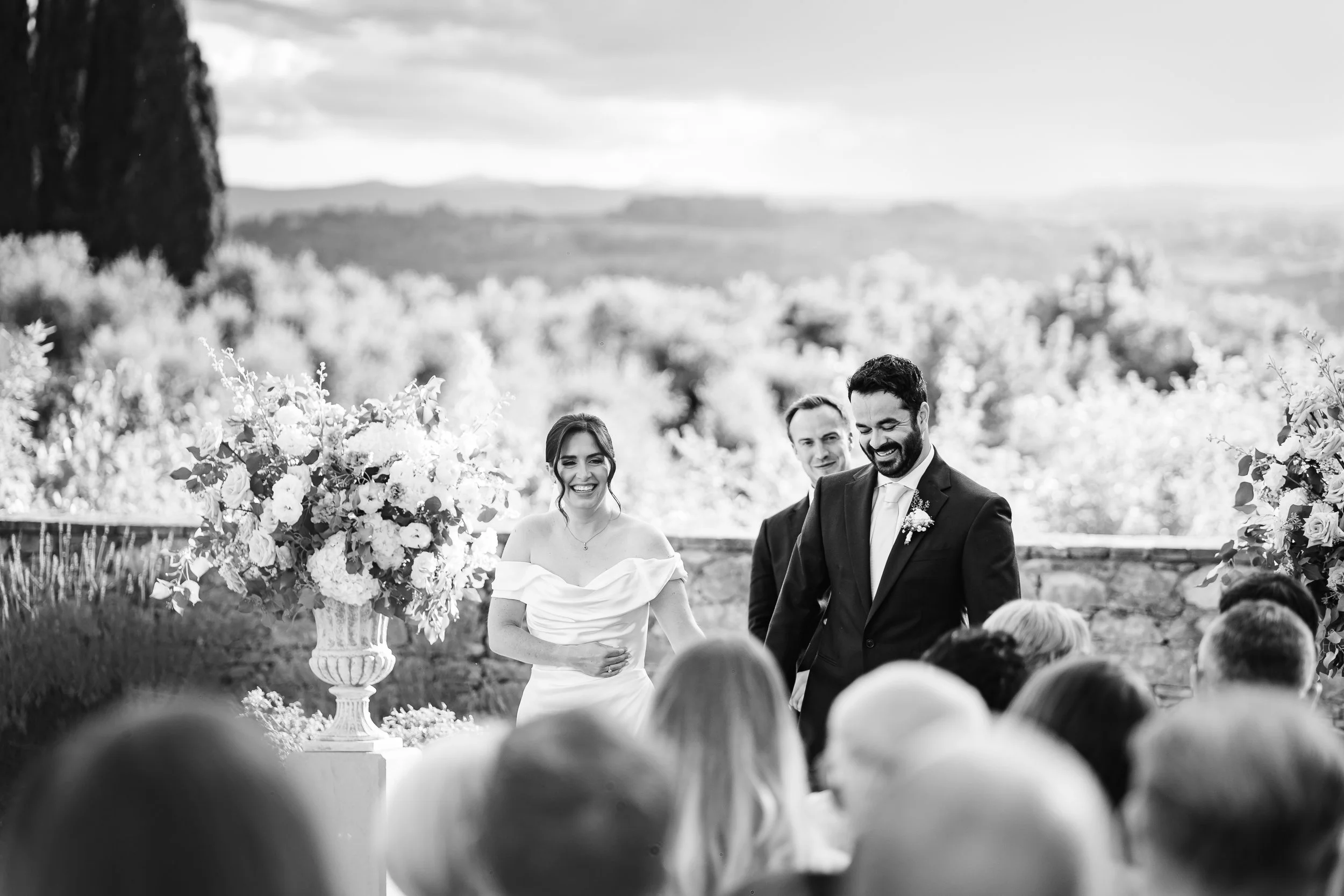 Foto in bianco e nero di una cerimonia di matrimonio all'aperto con il marito e la moglie sorridenti davanti a un pubblico, con un paesaggio naturale sullo sfondo, decorazioni floreali e un cielo nuvoloso.