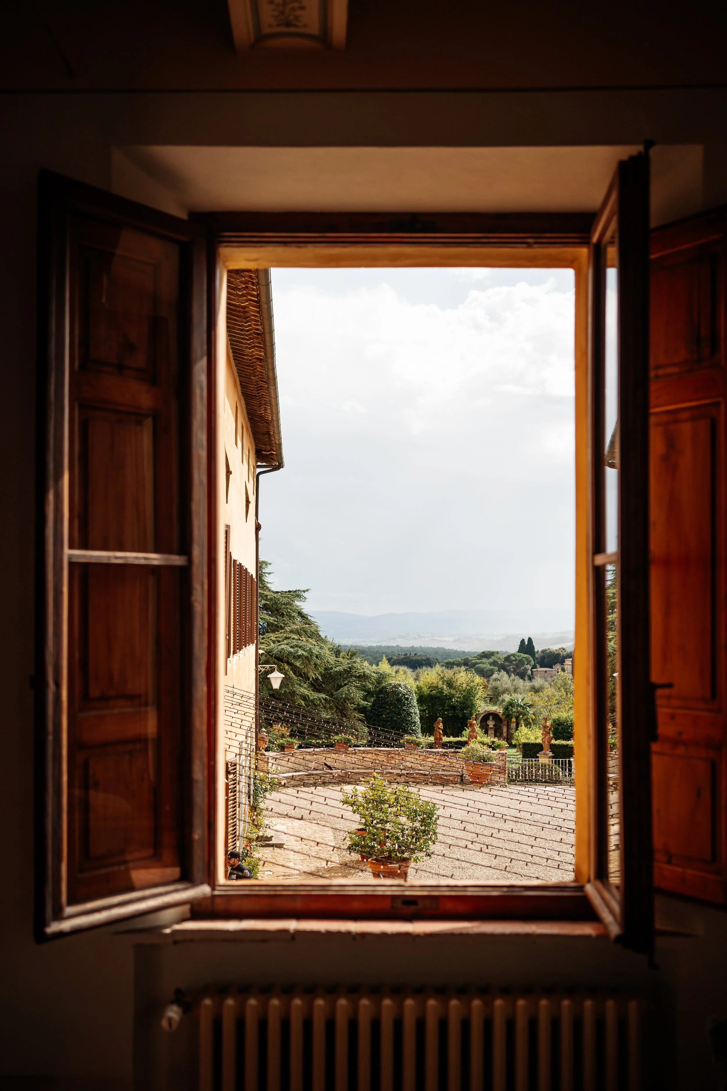 Vista da una finestra aperta che mostra un cortile rustico e paesaggi collinari lontani con nuvole nel cielo.