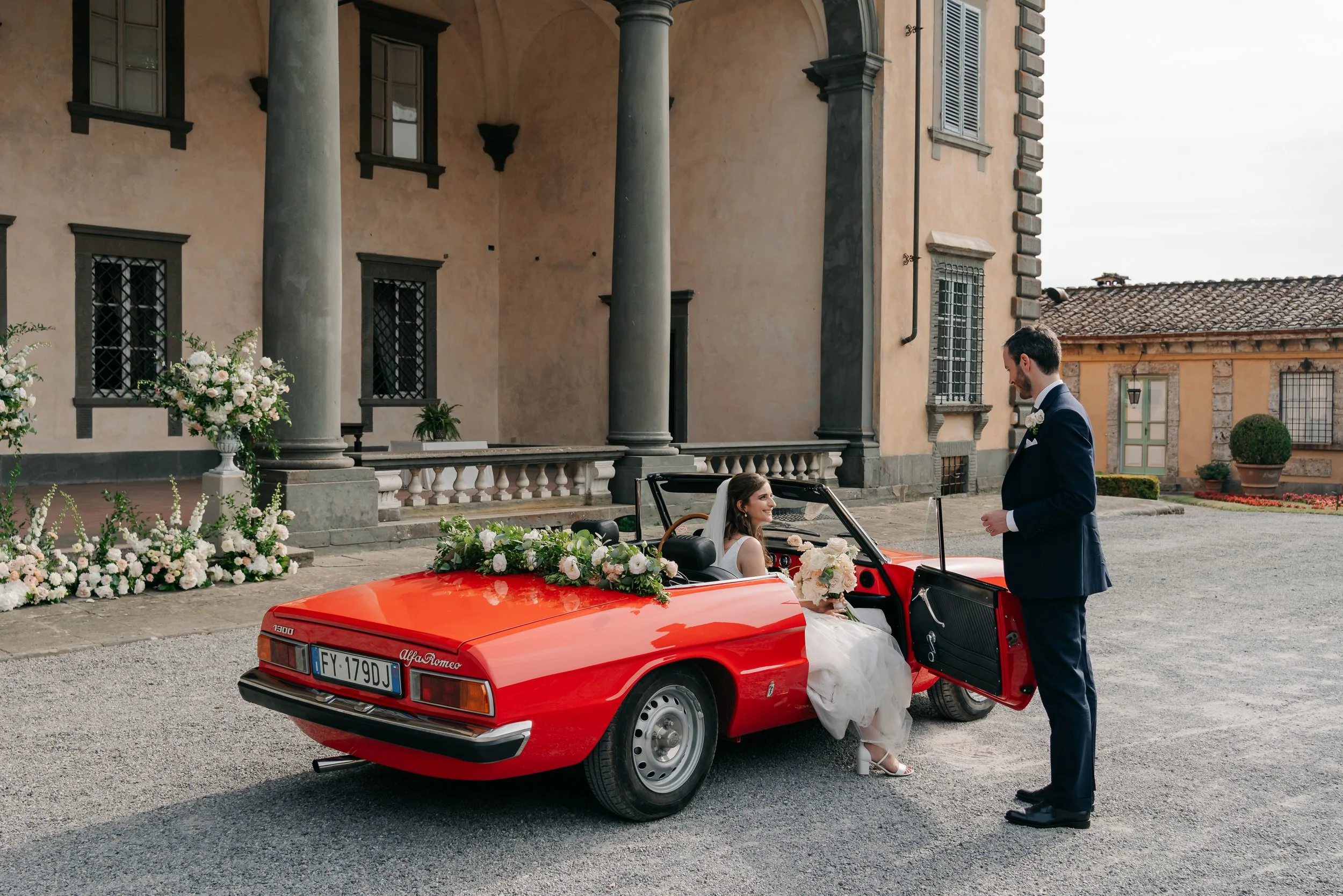 Una coppia di sposi, una donna in abito da sposa e un uomo in completo blu, interagiscono vicino a una macchina sportiva rossa decorata con fiori, davanti a un edificio storico.