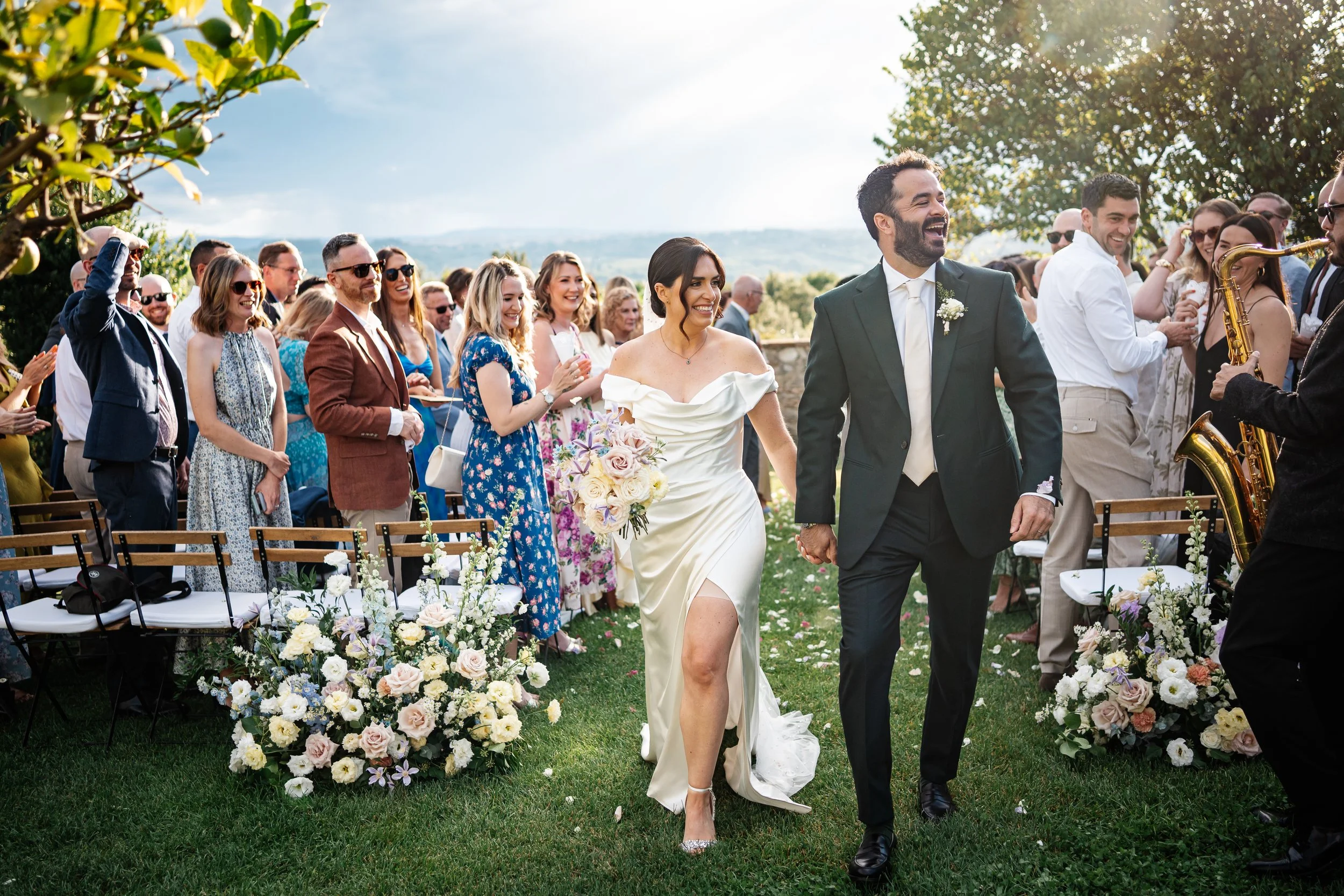 Coppia di sposi che cammina mano nella mano durante il matrimonio all'aperto, circondata da amici e parenti che applaudono e sorridono, con decorazioni floreali e un cielo soleggiato.