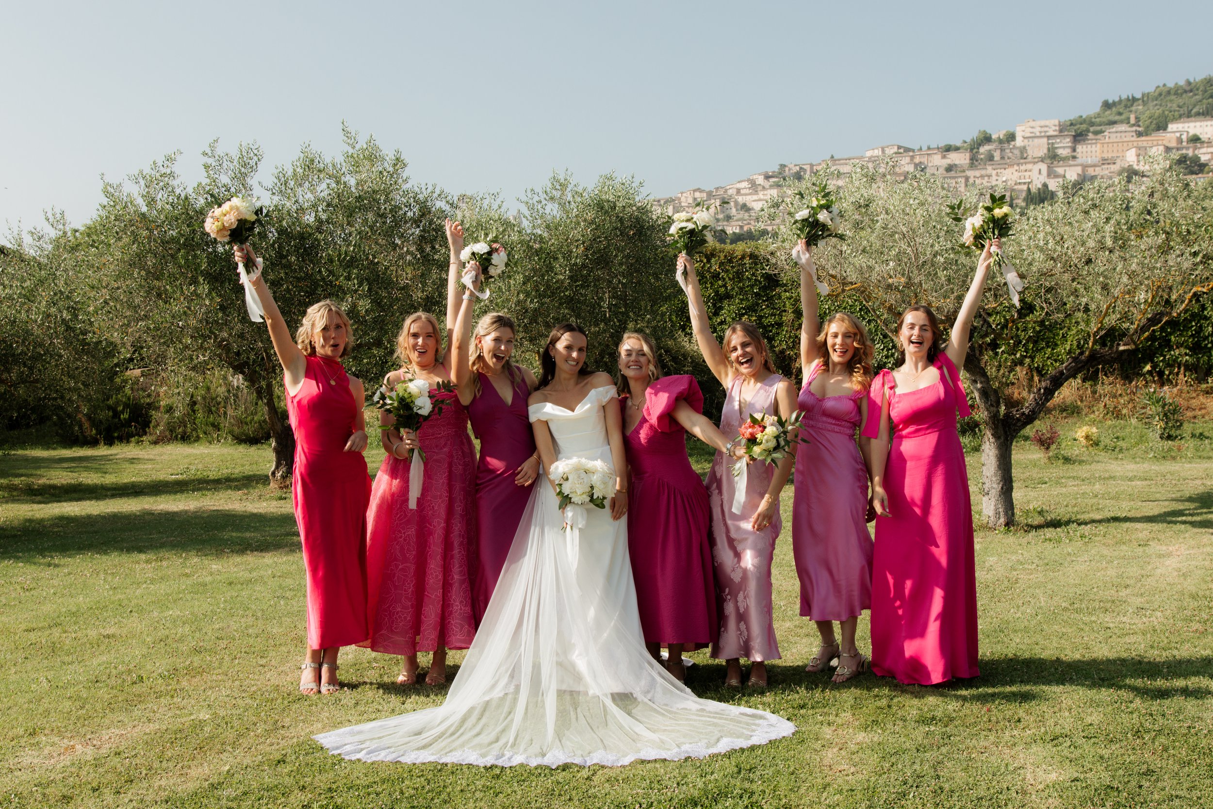 Gruppo di donne sorridenti in abiti da sera colorati, probabilmente durante un matrimonio, che tengono bouquet di fiori, in un giardino con alberi e un paesaggio collinare sullo sfondo.