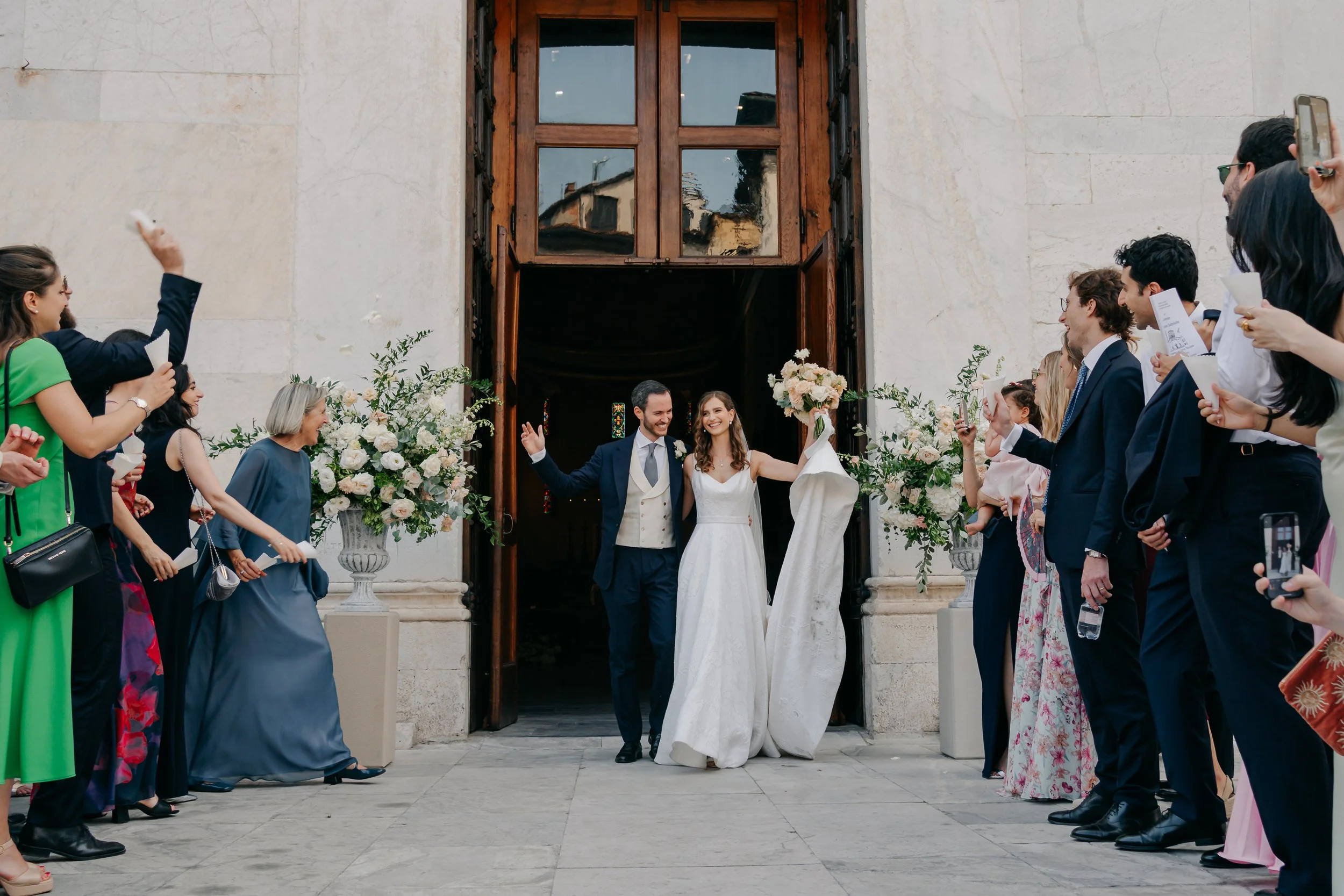 Una coppia di sposi entra in chiesa circondata da amici e parenti che scattano foto e li salutano, decorazioni floreali e un portale in legno dietro di loro.