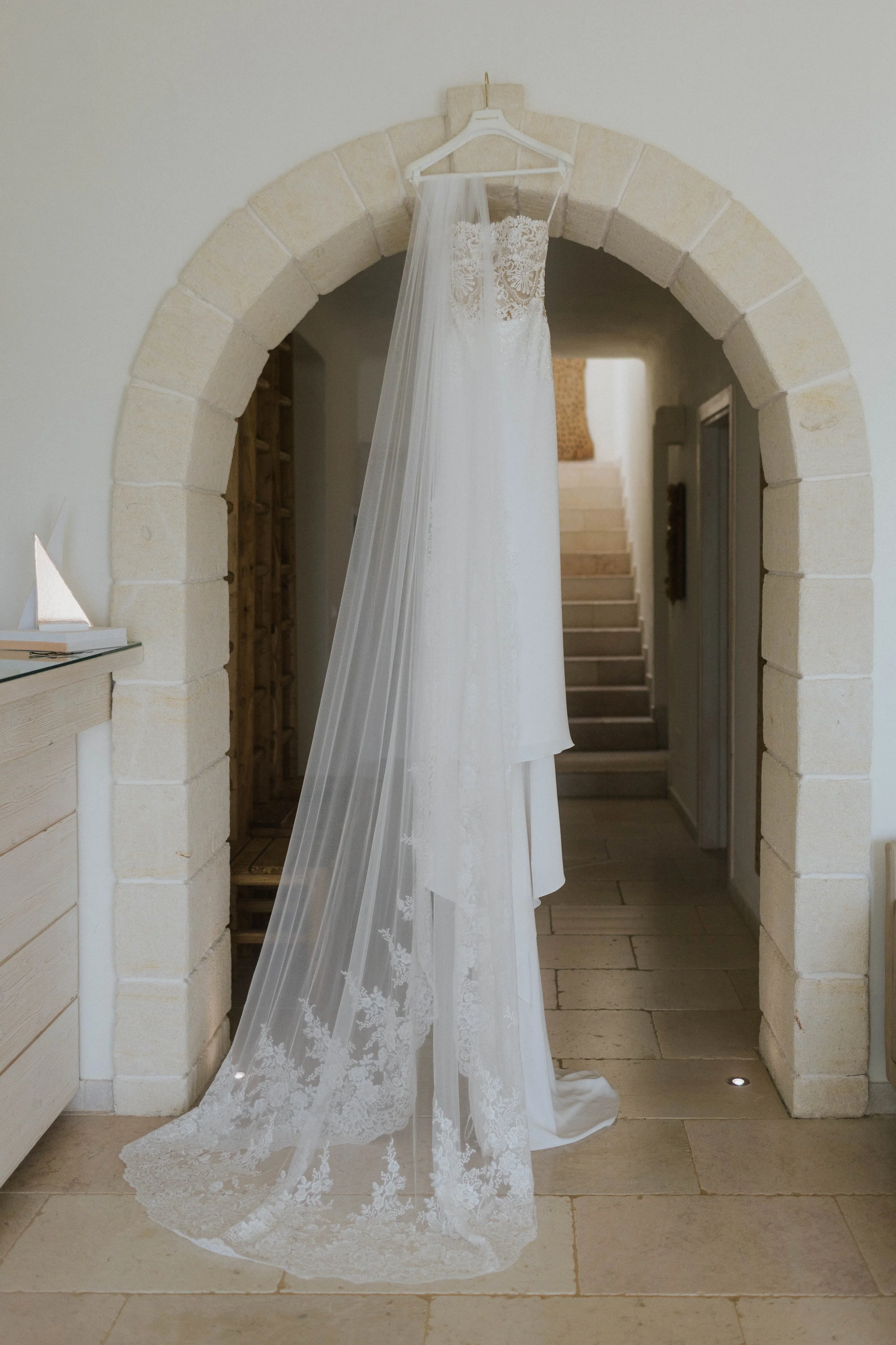 Abito da sposa bianco con velo ricamato appeso a uno scivolo davanti a un arco di pietra.