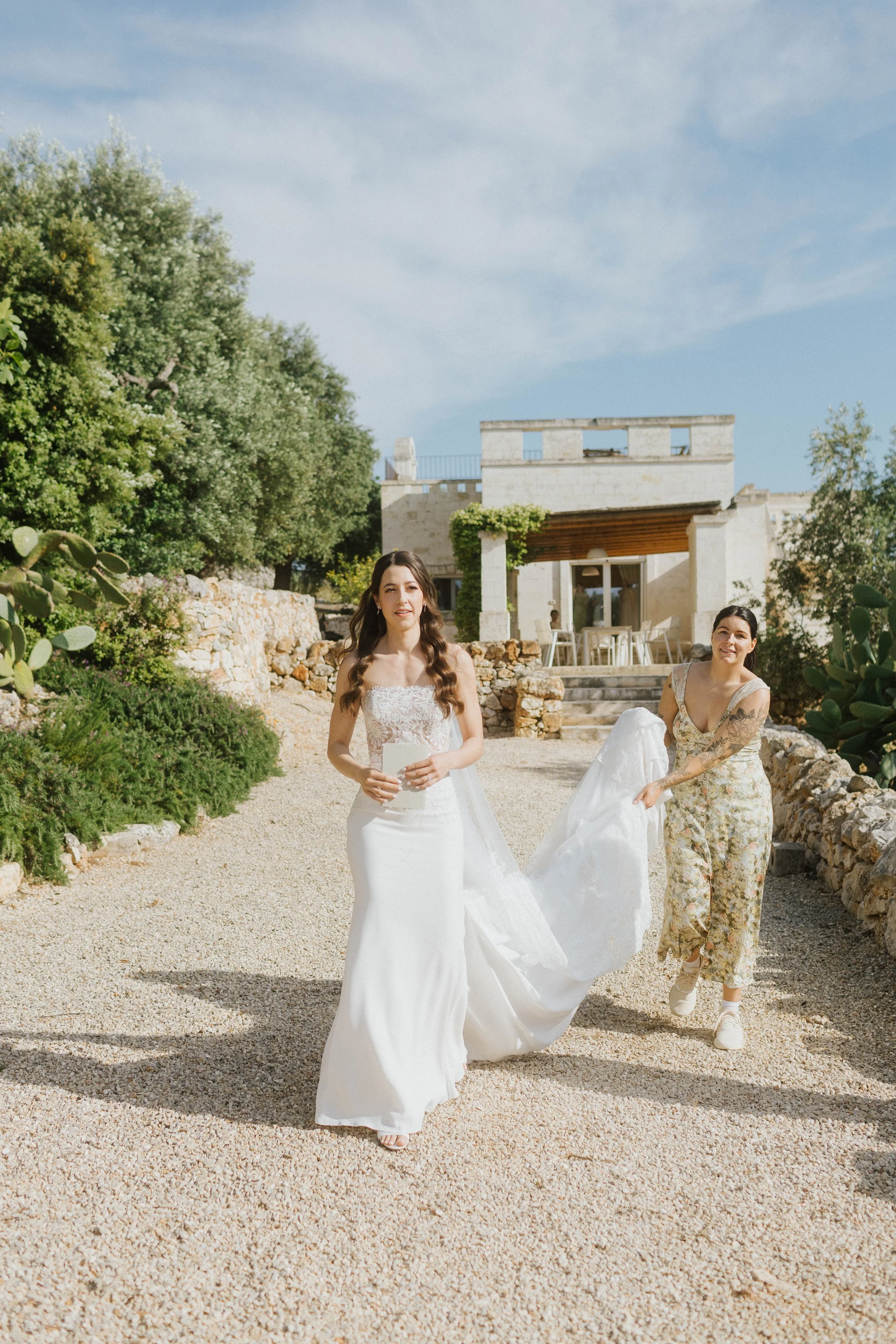 Una coppia di donne con abiti da sposa che camminano all'aperto in un giardino soleggiato.