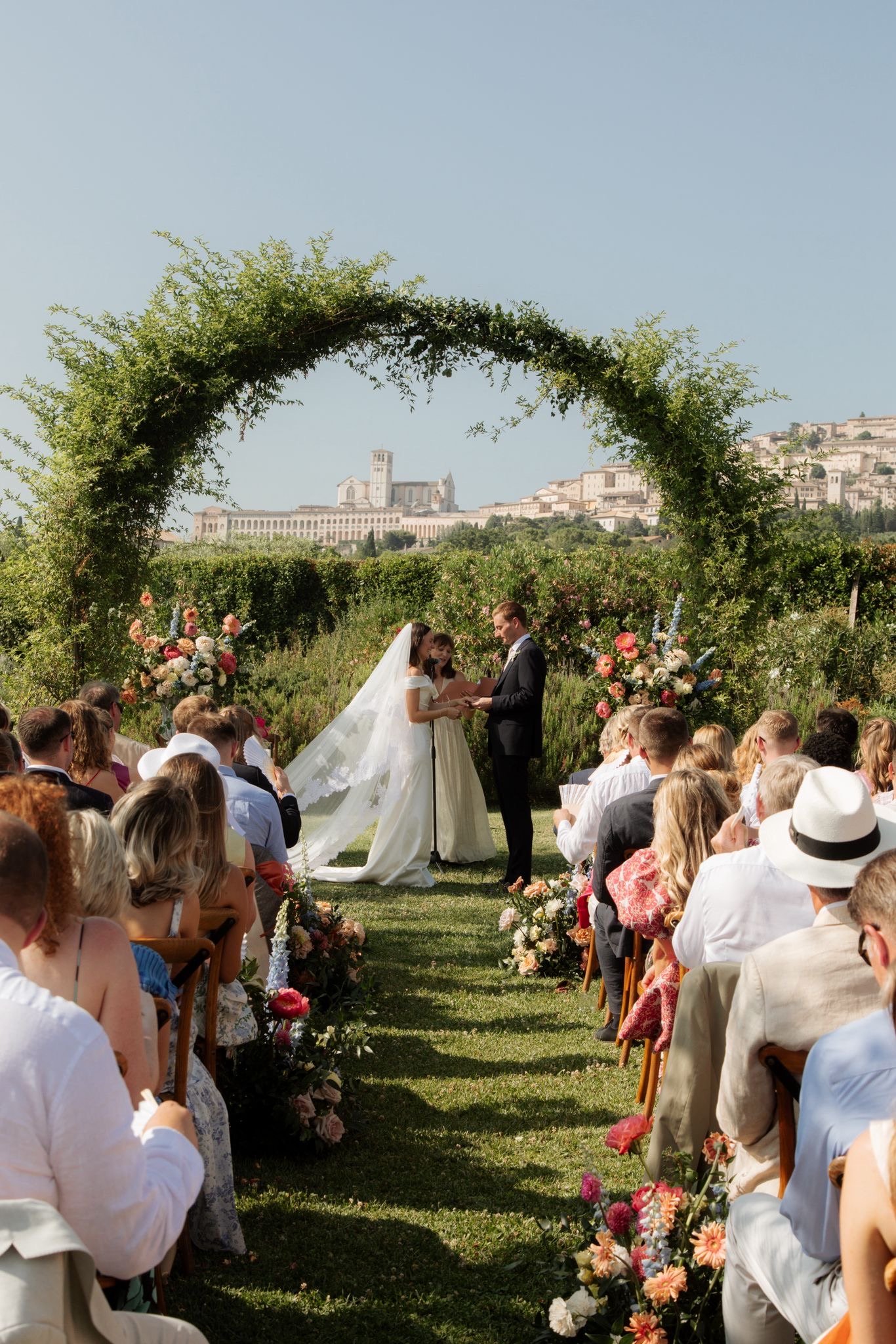 Coppia di sposi durante il loro matrimonio sotto un arco decorato con fiori, con invitati seduti ai lati e un castello sullo sfondo in un paesaggio collinare
