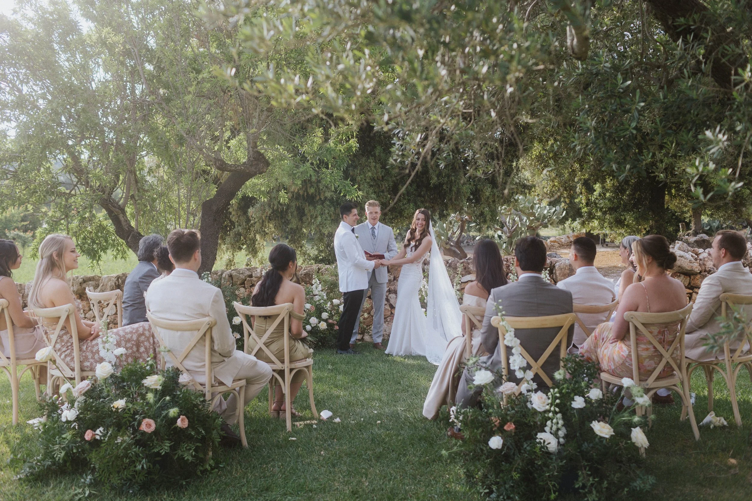 Una cerimonia di matrimonio all'aperto sotto un grande albero con gli sposi e ospiti seduti su sedie di legno, decorazioni floreali e un ambiente naturale.