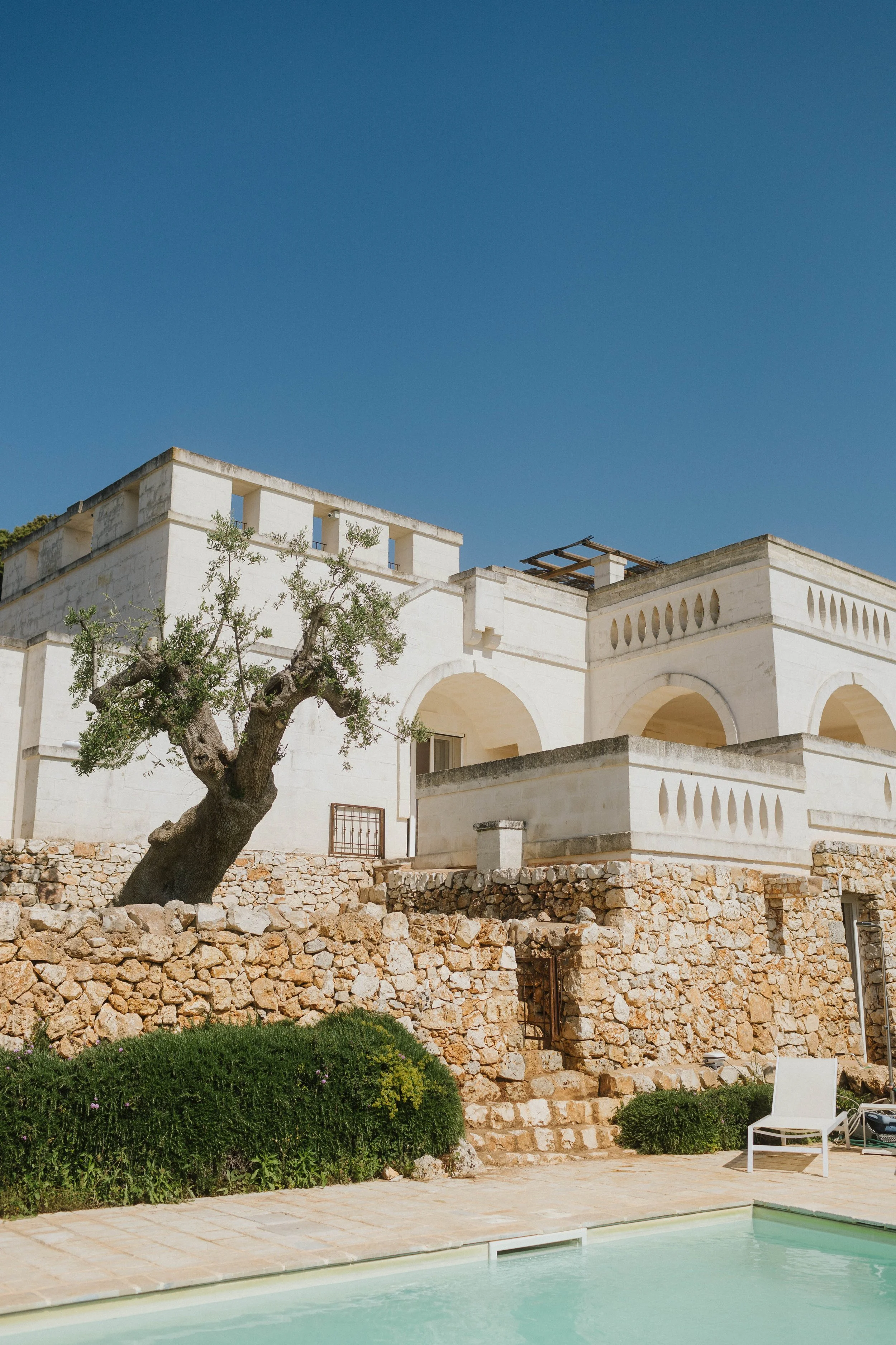Casa in stile mediterraneo con muri bianchi, arco e mura in pietra, alberi e piscina in primo piano, cielo azzurro.