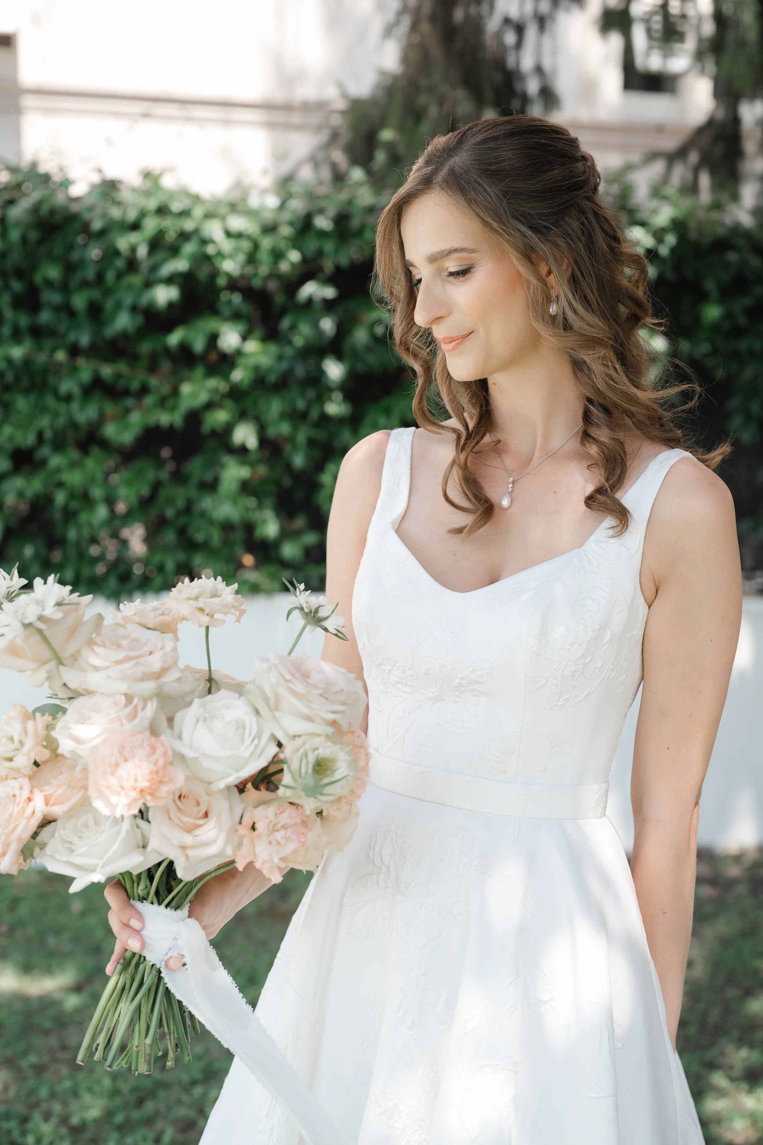 Una donna in abito da sposa bianco tiene un mazzo di fiori rosa e crema, ambientato all'aperto con uno sfondo di cespugli verdi.