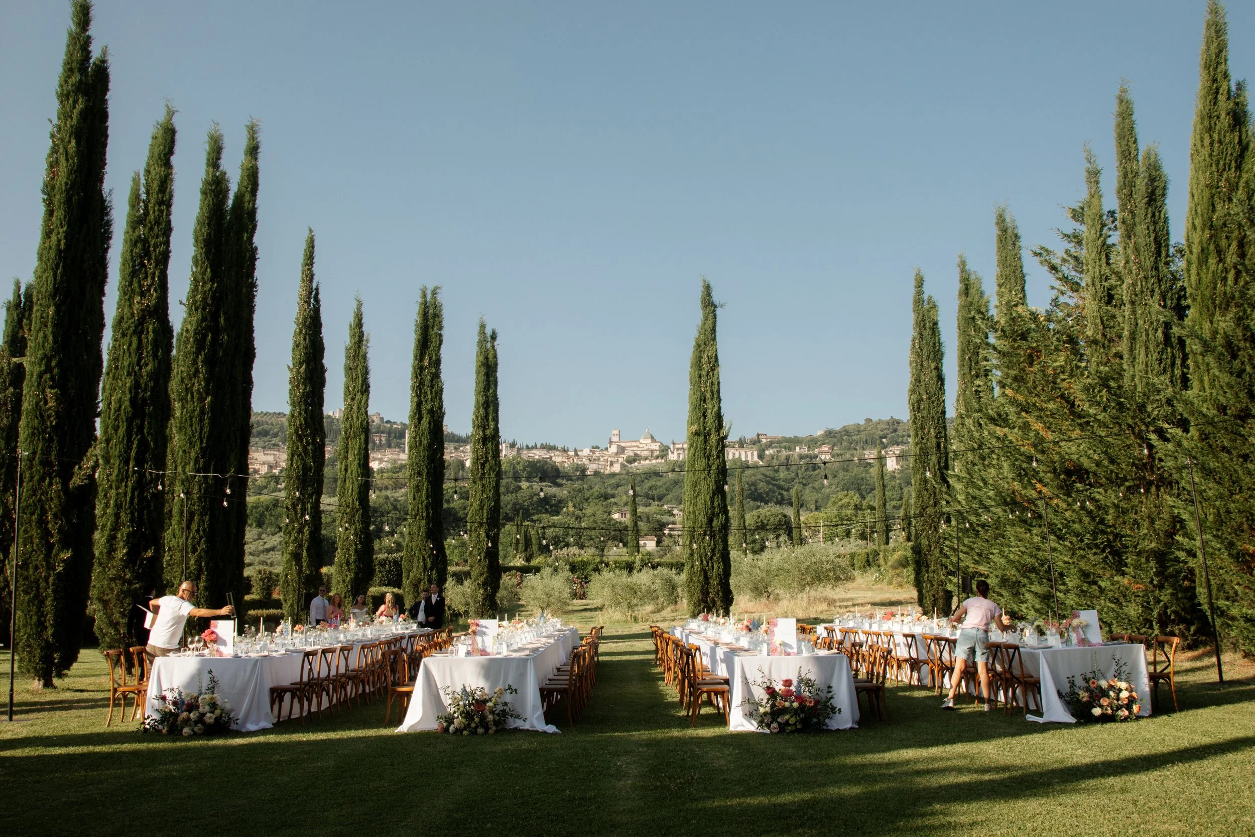 Tavola apparecchiata all'aperto con dettagli floreali, in un grande giardino con filari di cipressi e un panorama collinare in background.