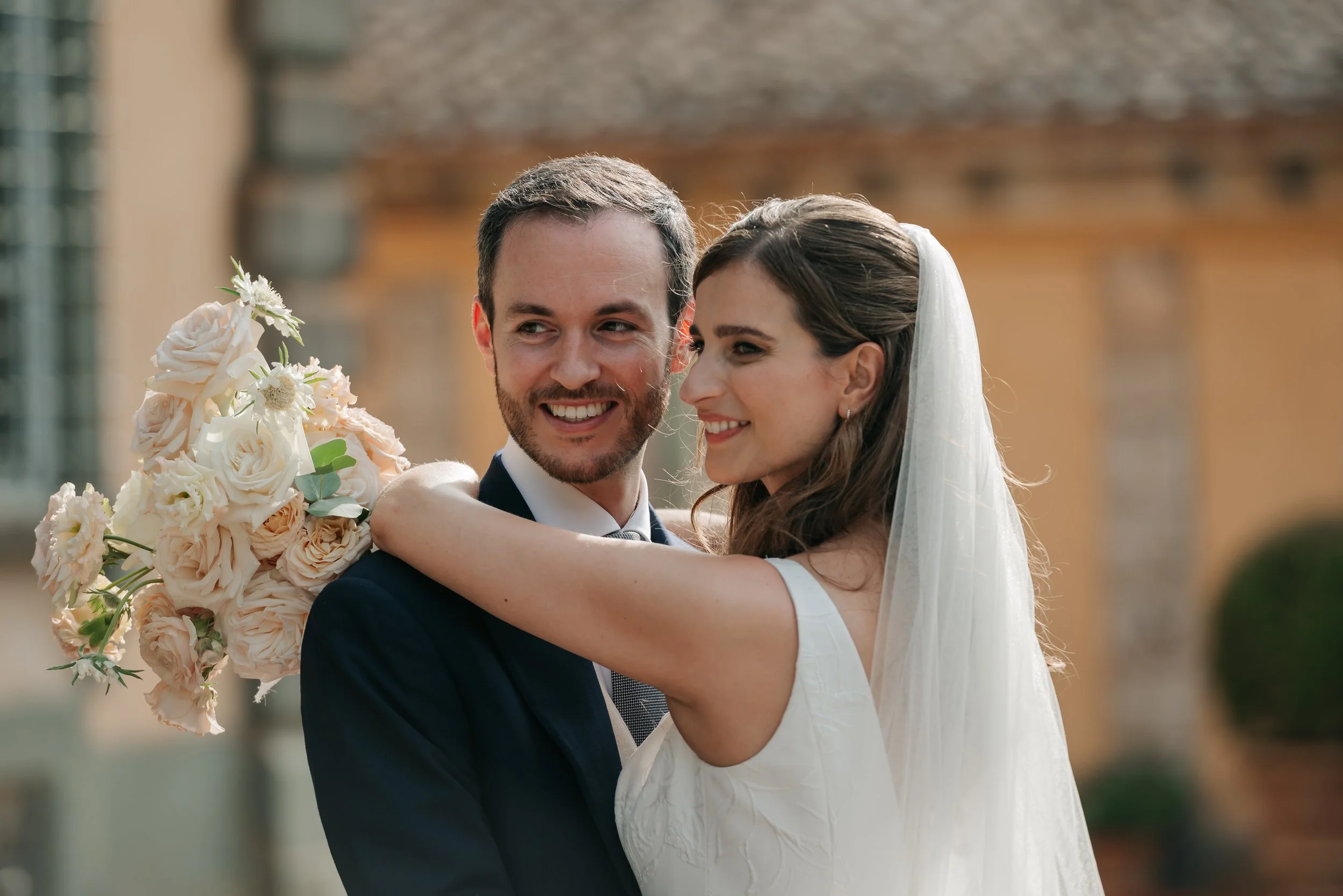 Una coppia di sposi sorridenti si abbraccia, con il marito che indossa un abito blu scuro e la sposa un vestito bianco con velo, mentre lei tiene un bouquet di fiori color rosa chiaro, sullo sfondo di un edificio antico.