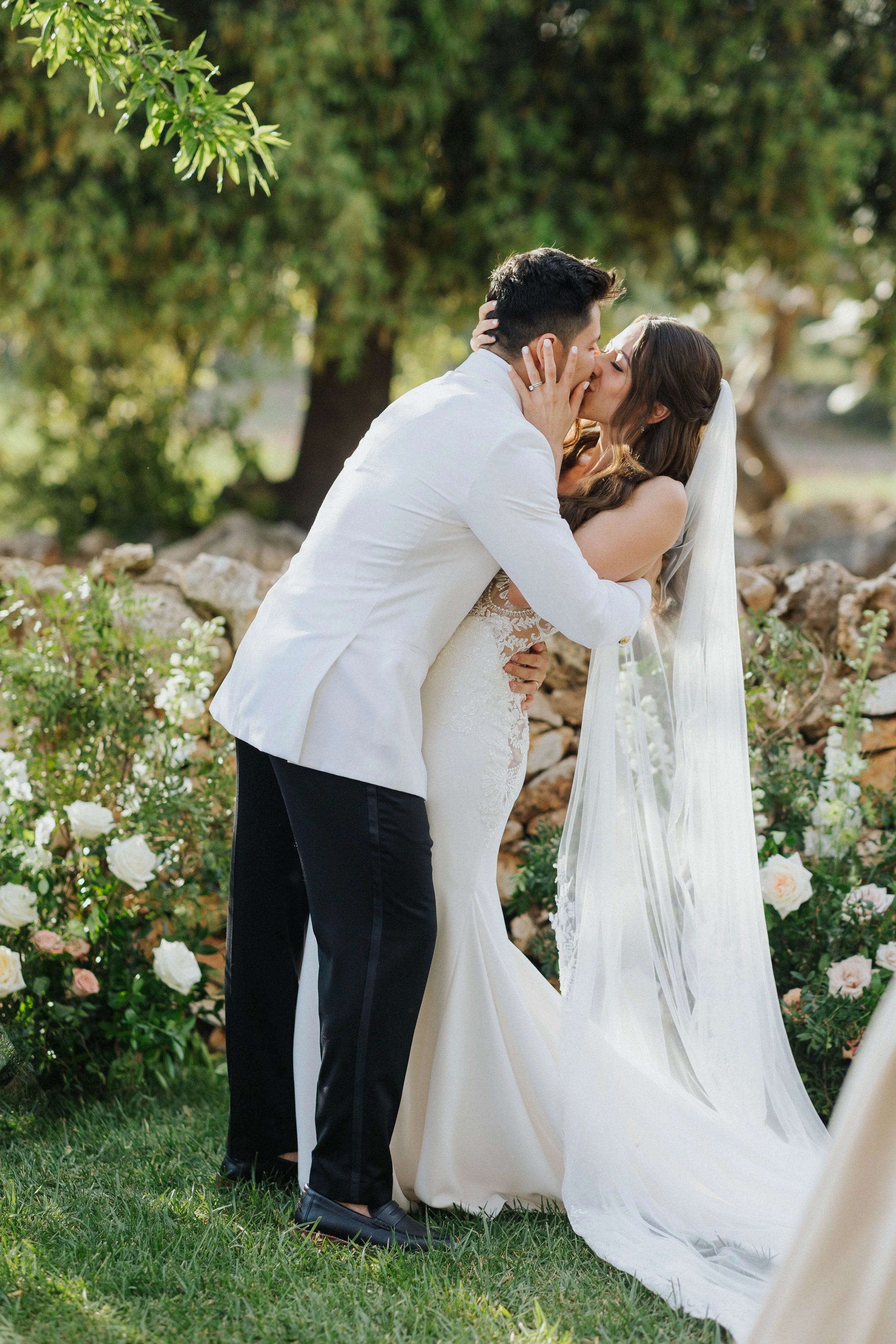 Una coppia di sposi si bacia durante il matrimonio all'aperto, circondati da fiori e vegetazione.