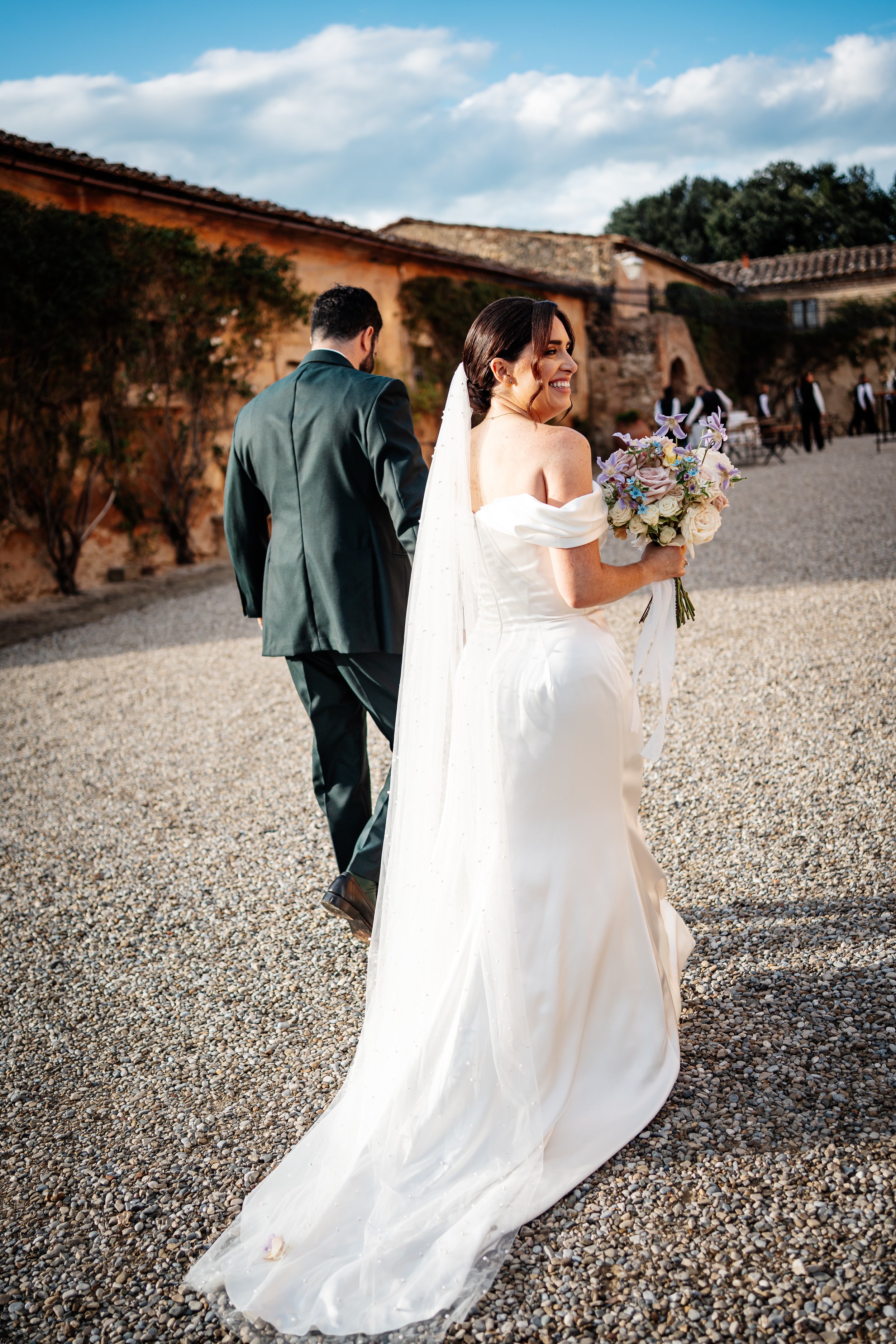 Una sposa con un abito bianco e un velo sorridente, che tiene un bouquet di fiori, cammina su un piazzale di ciottoli durante il tramonto, con uno sfondo di edifici rustici e cielo nuvoloso.