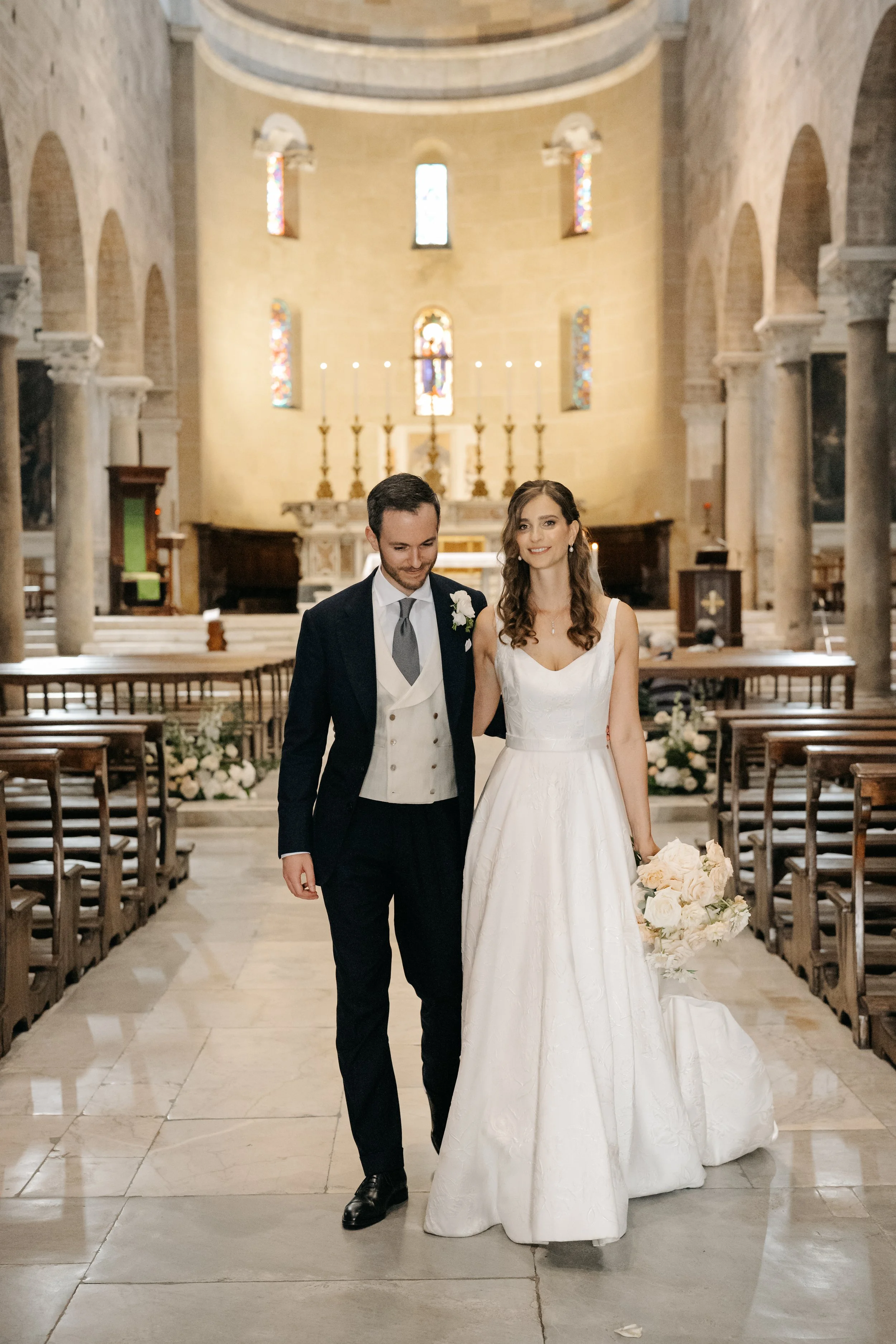 Sposo e sposa che camminano all'interno di una chiesa, atmosfera di matrimonio, decorazioni floreali e altare in background.