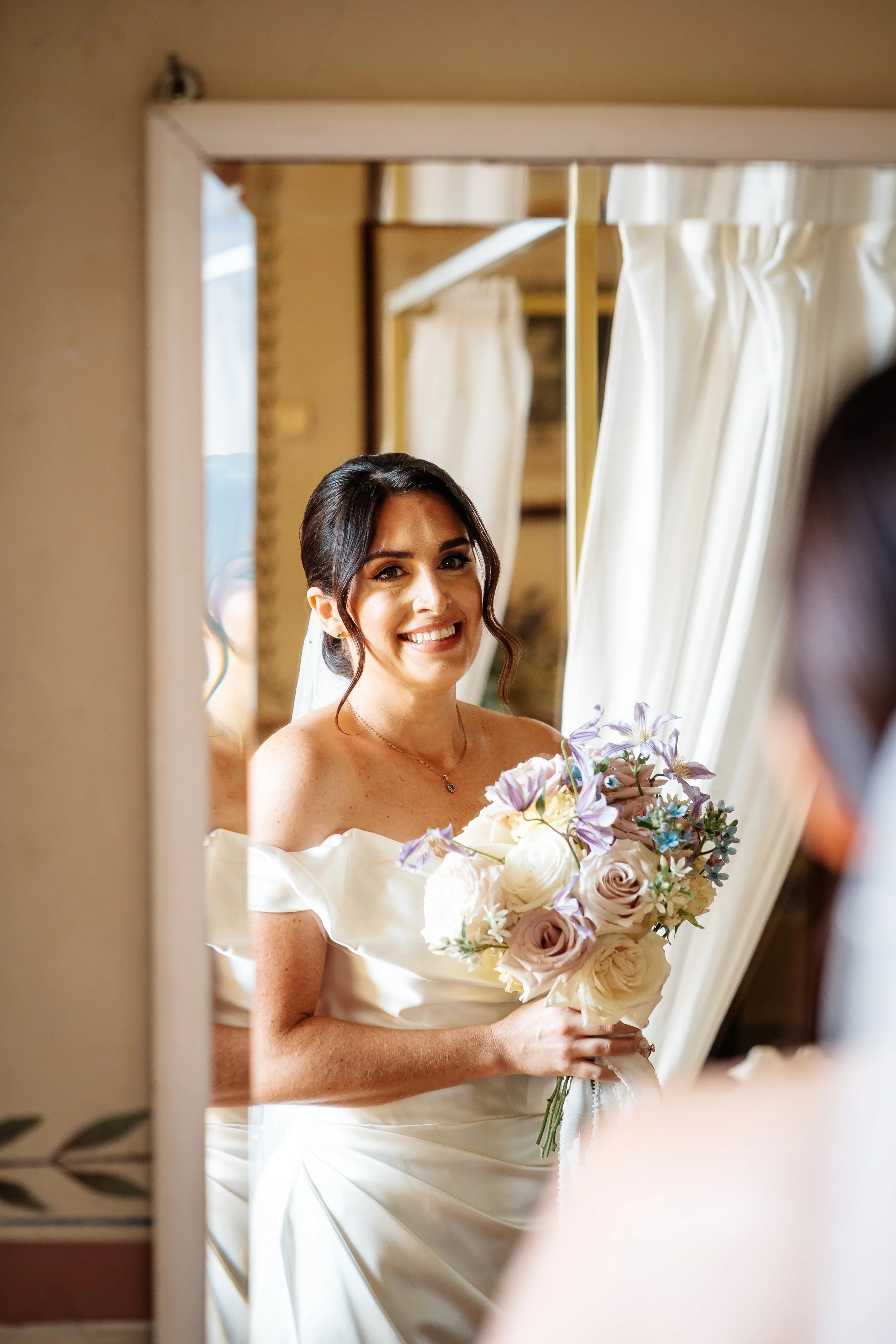 Sposa in abito bianco che si guarda nello specchio, sorride e tiene un bouquet di fiori.