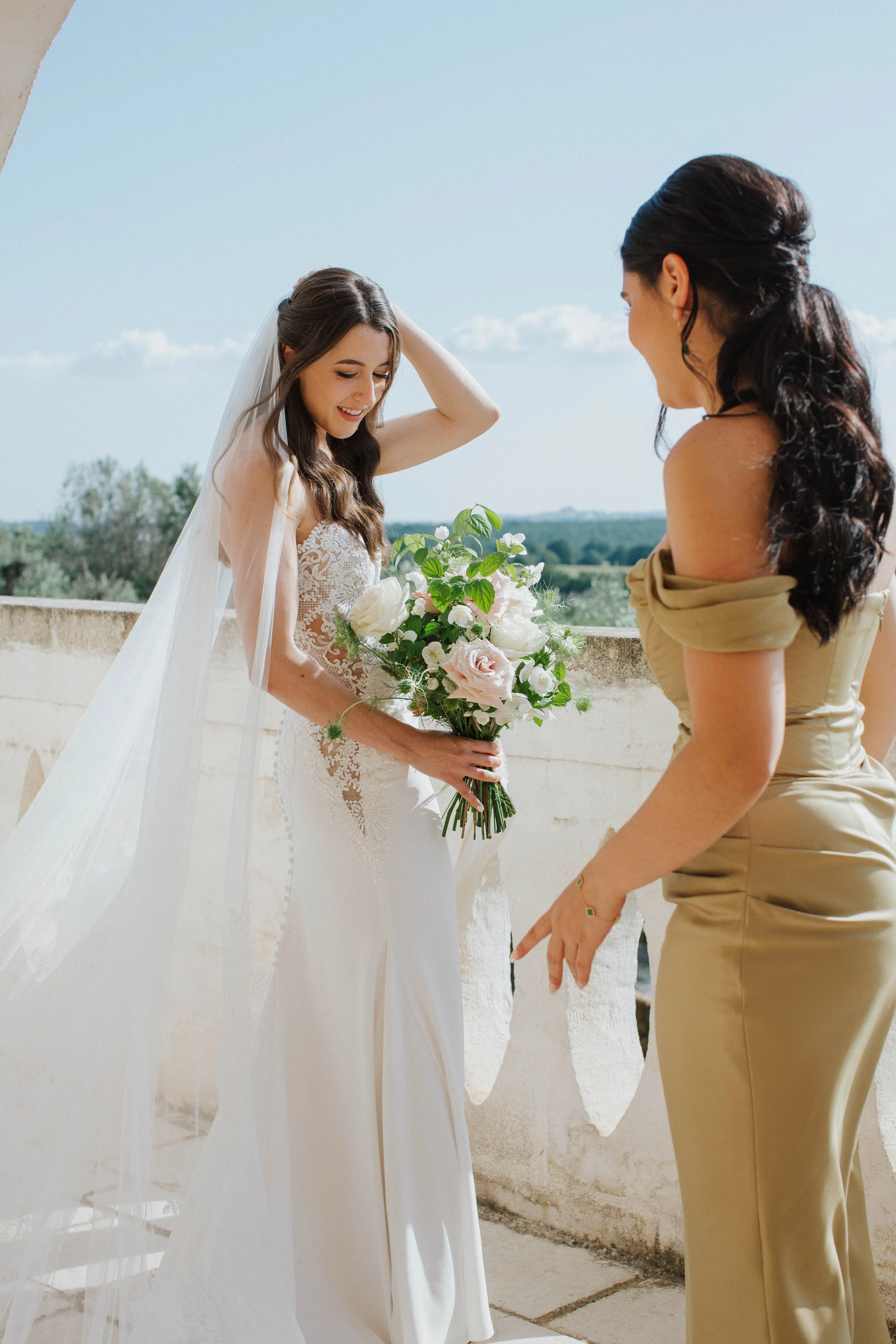 Sposa in abito bianco con bouquet di fiori, assistita da donna in abito beige, sul terrazzo con vista panoramica.
