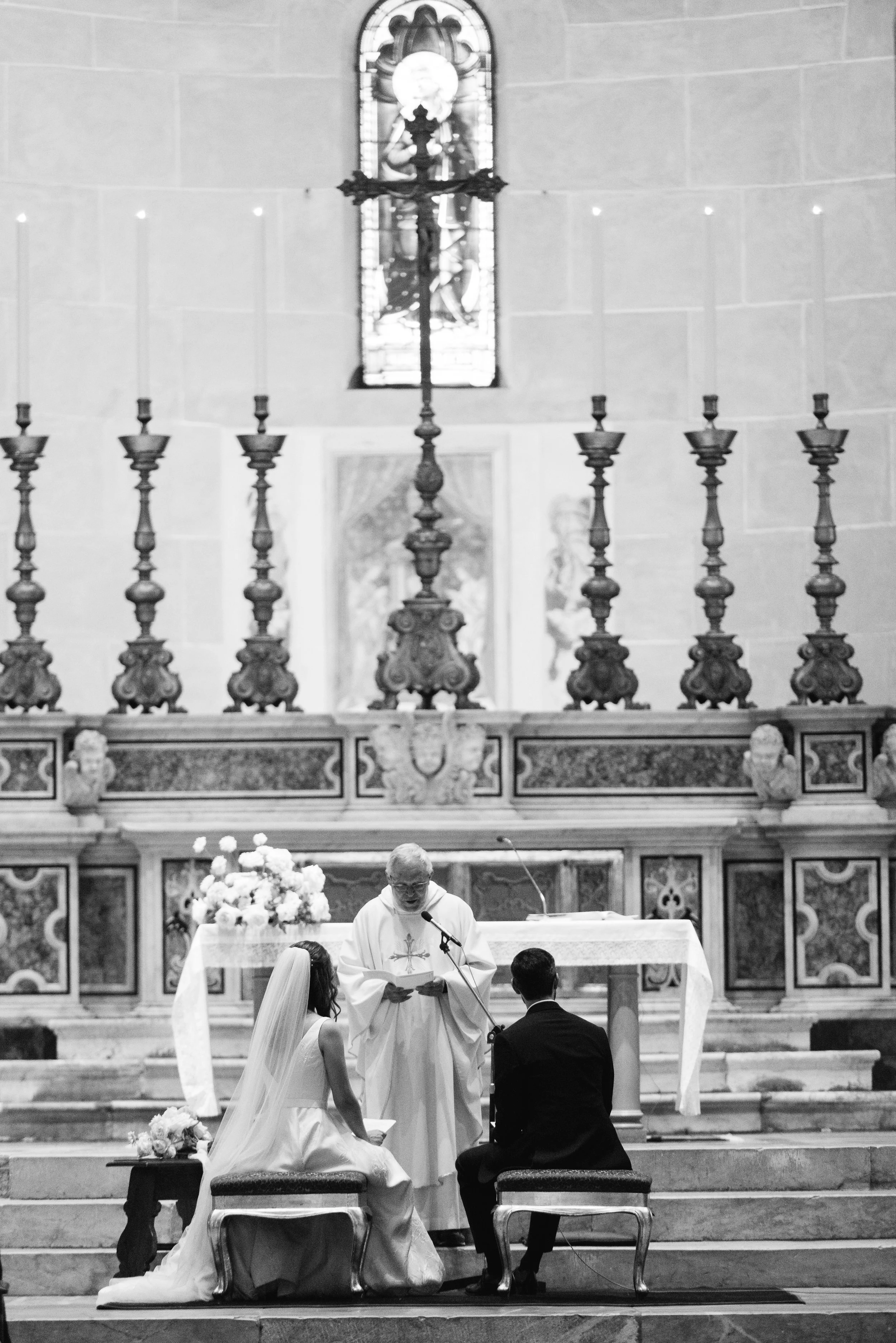 Matimonio in chiesa, sacerdote, sposi seduti durante la cerimonia, decorazioni floreali, atmosfera religiosa.