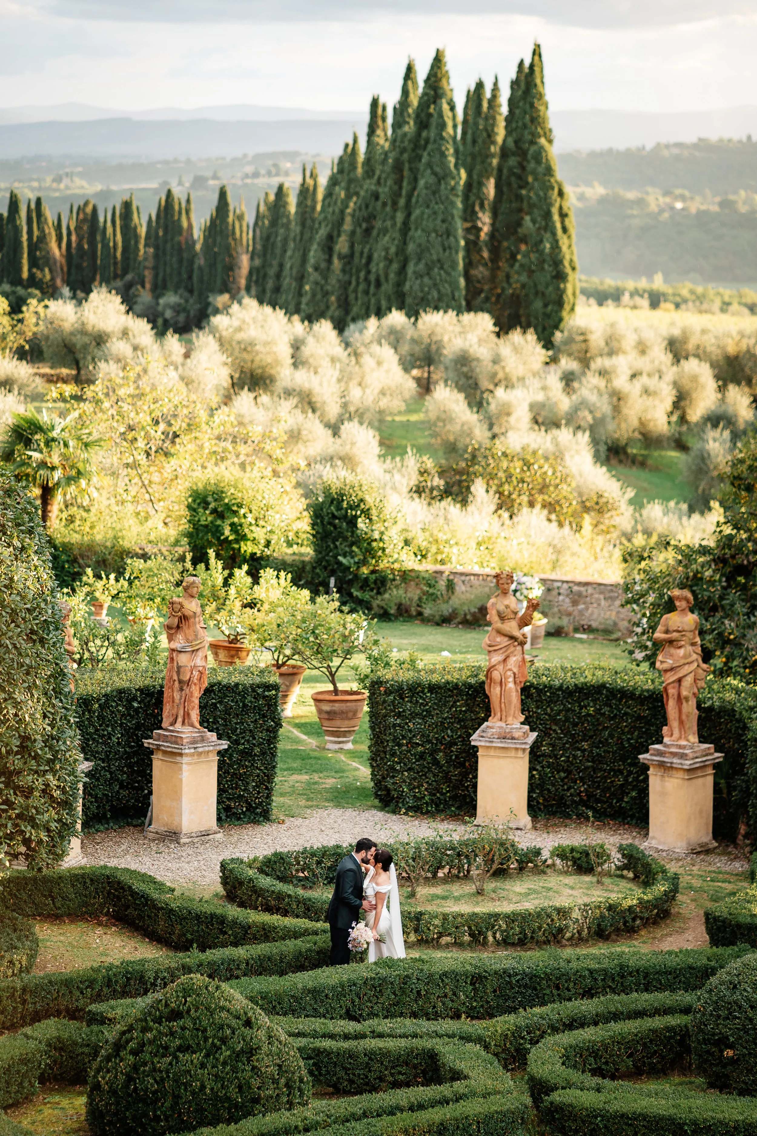 Una coppia di sposi si bacia in un giardino con statue antiche e piante ornamentali, con un paesaggio collinare e alberi alti sullo sfondo.